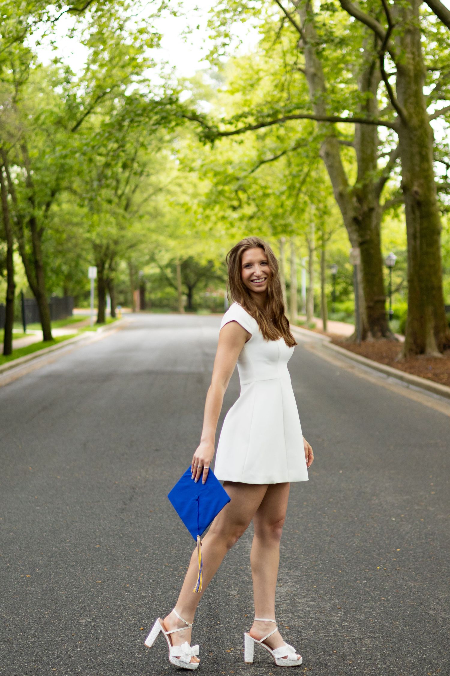 Graduate in white dress and blue robe poses confidently on tree-lined street.
 at the University of Delaware.