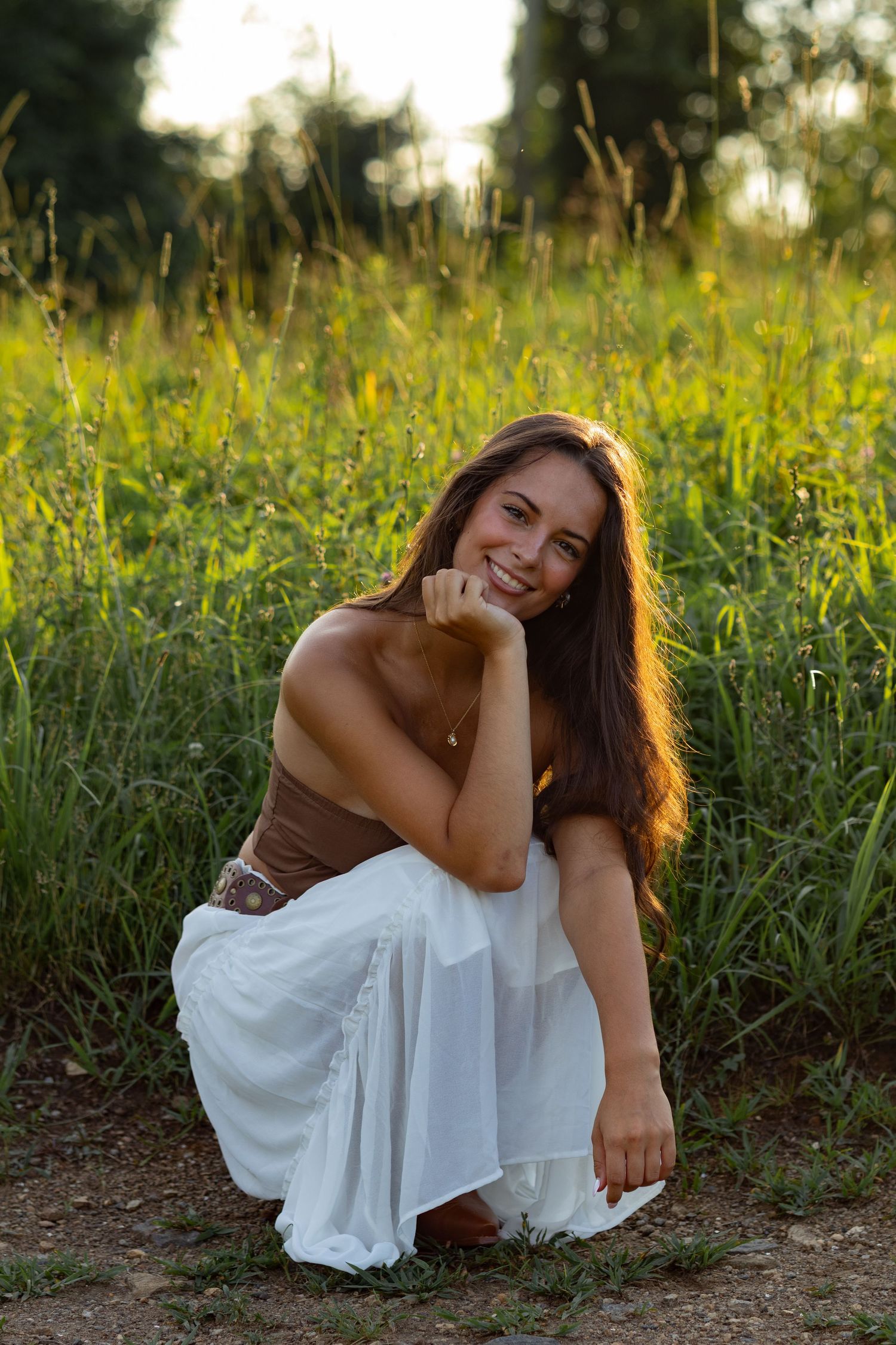 Portrait in brown top and flowing white skirt crouching in summer meadow at sunset in Wilmington, Delaware.