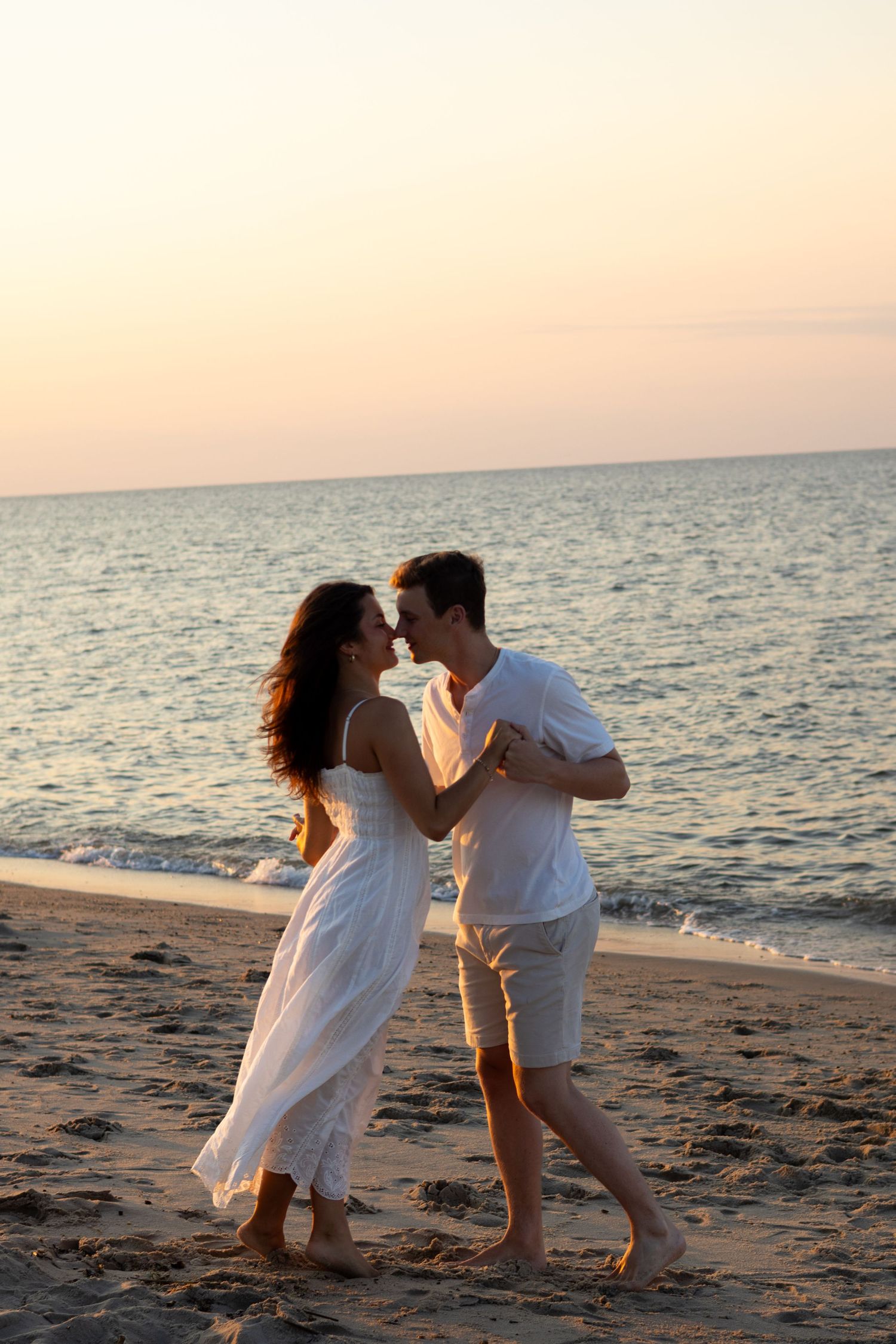 A couple in white clothing dances playfully on the beach with the ocean horizon behind them for engagement photos in Lewes, Delaware.