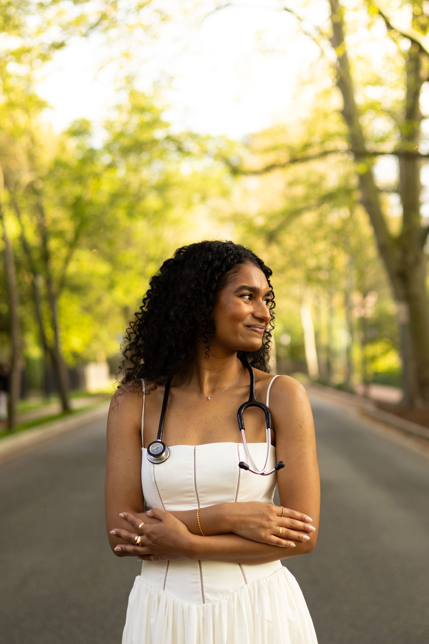 A person in a white dress stands thoughtfully on a tree-lined path during golden hour at the University of Delaware.