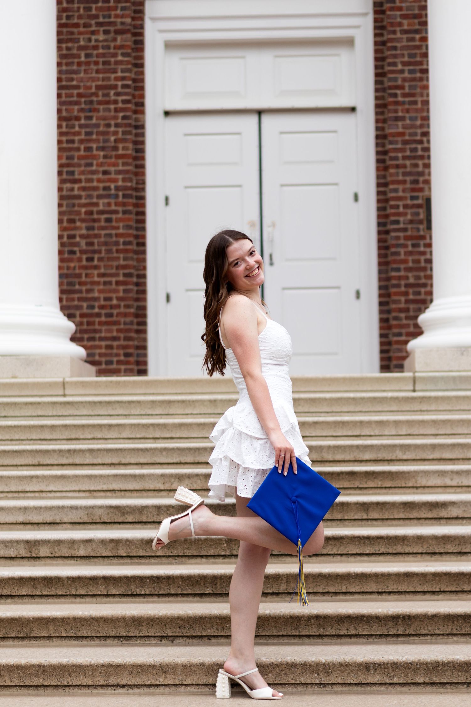 Graduate in white dress poses on brick steps with white columns at the University of Delaware.