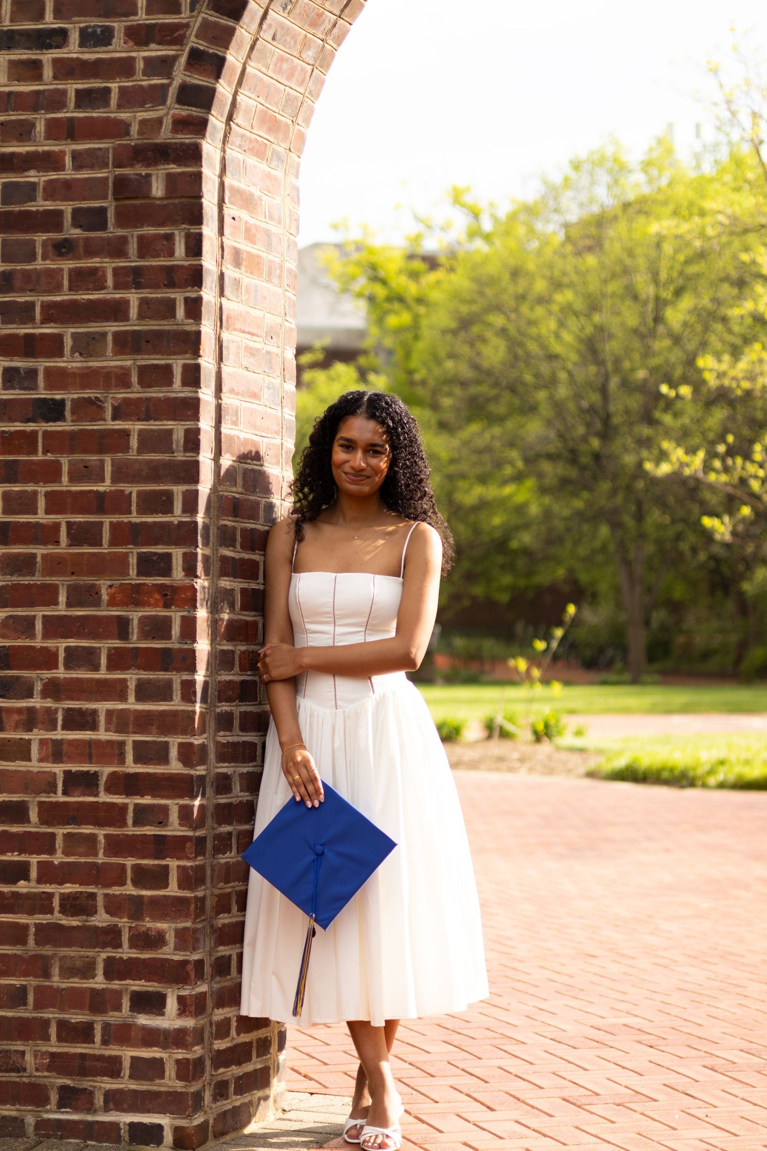 A person in a white dress poses with a blue graduation cap near a brick archway at the University of Delaware.