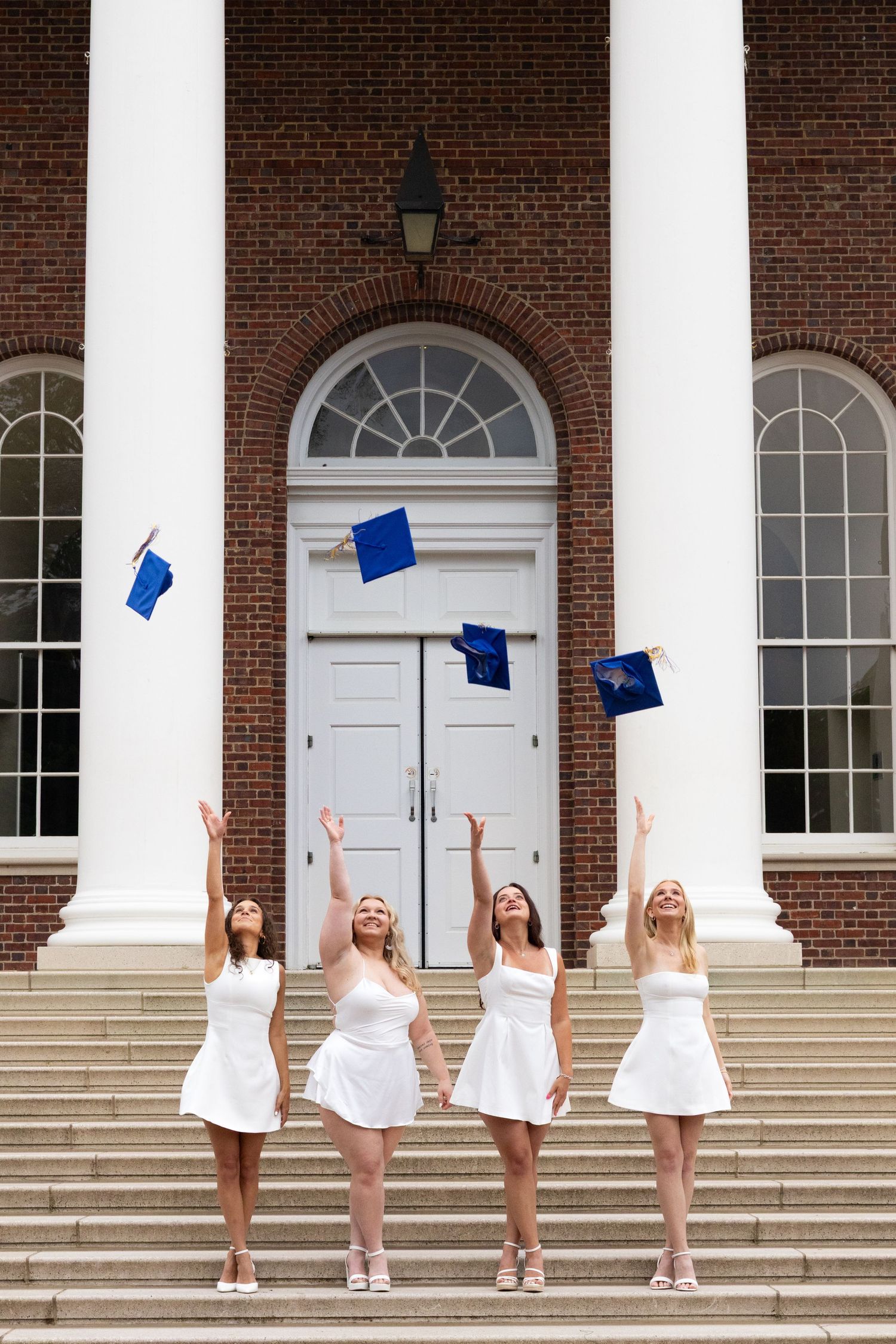 Graduates in white dresses toss blue graduation caps in front of columned building at the University of Delaware.