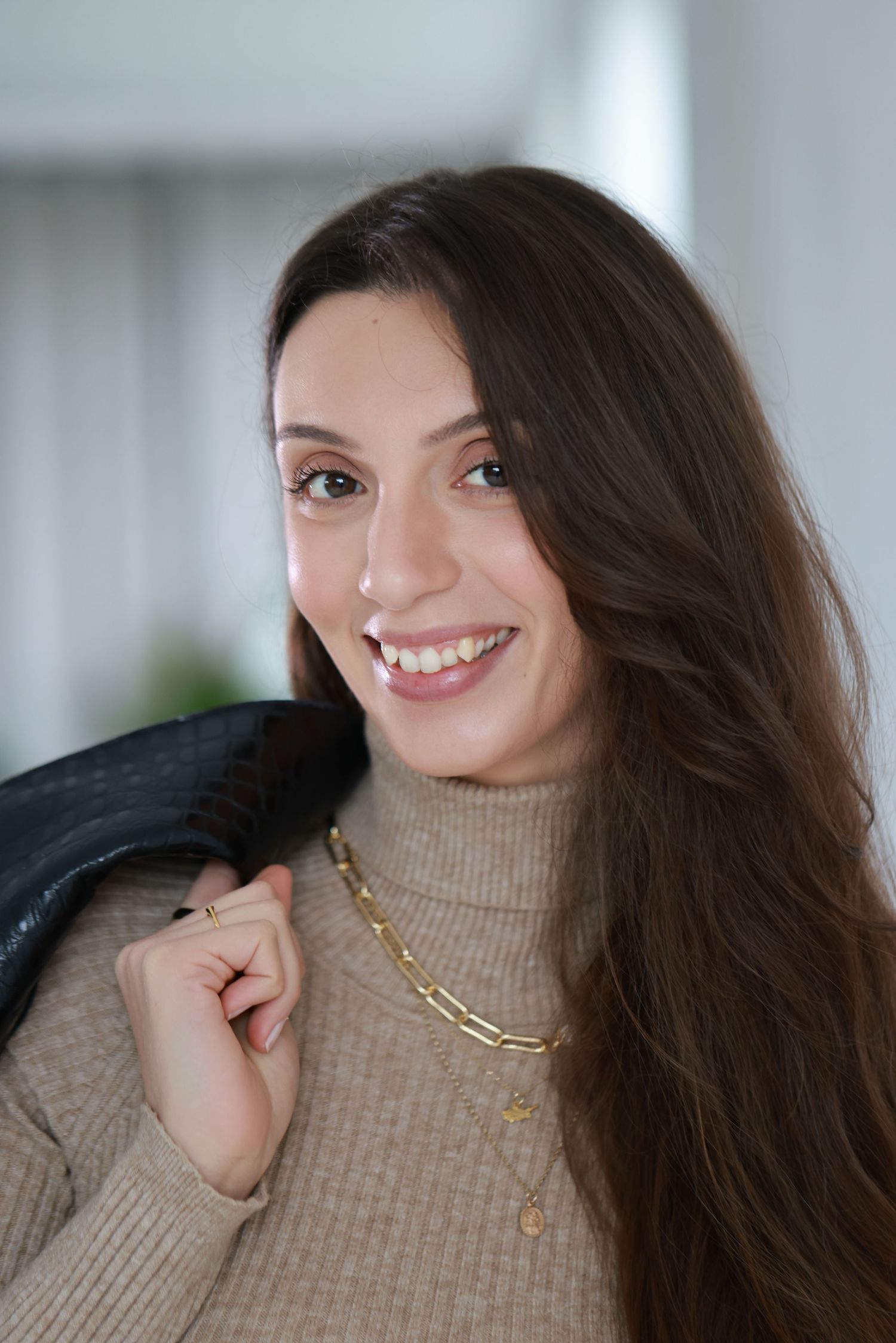 A person with long dark hair and a beige turtleneck sweater smiles warmly while wearing a gold chain necklace.
