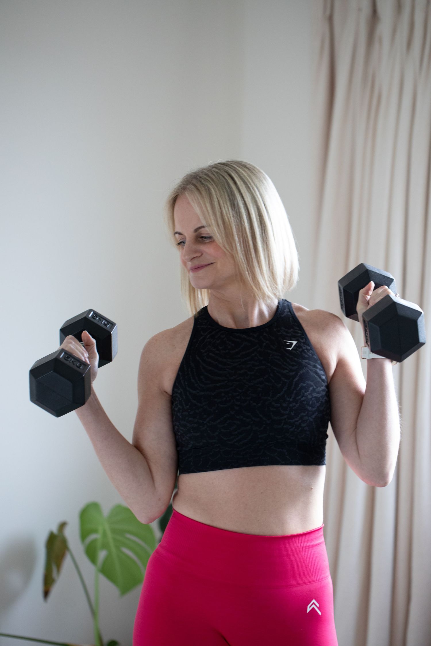Person in black workout top and pink leggings demonstrates fitness poses against white wall with houseplant.