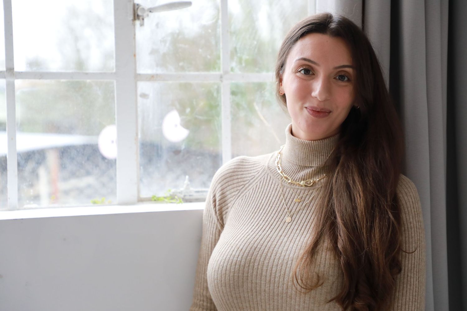 A woman in a beige turtleneck sweater poses near a bright window in a casual indoor setting.