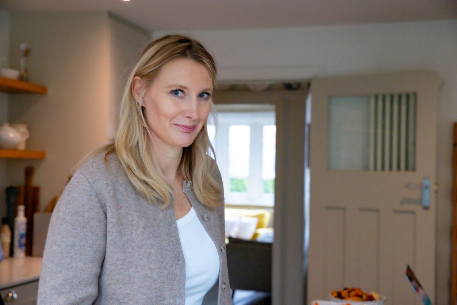 Woman in a gray cardigan and white top stands in a modern home interior with wooden shelves.