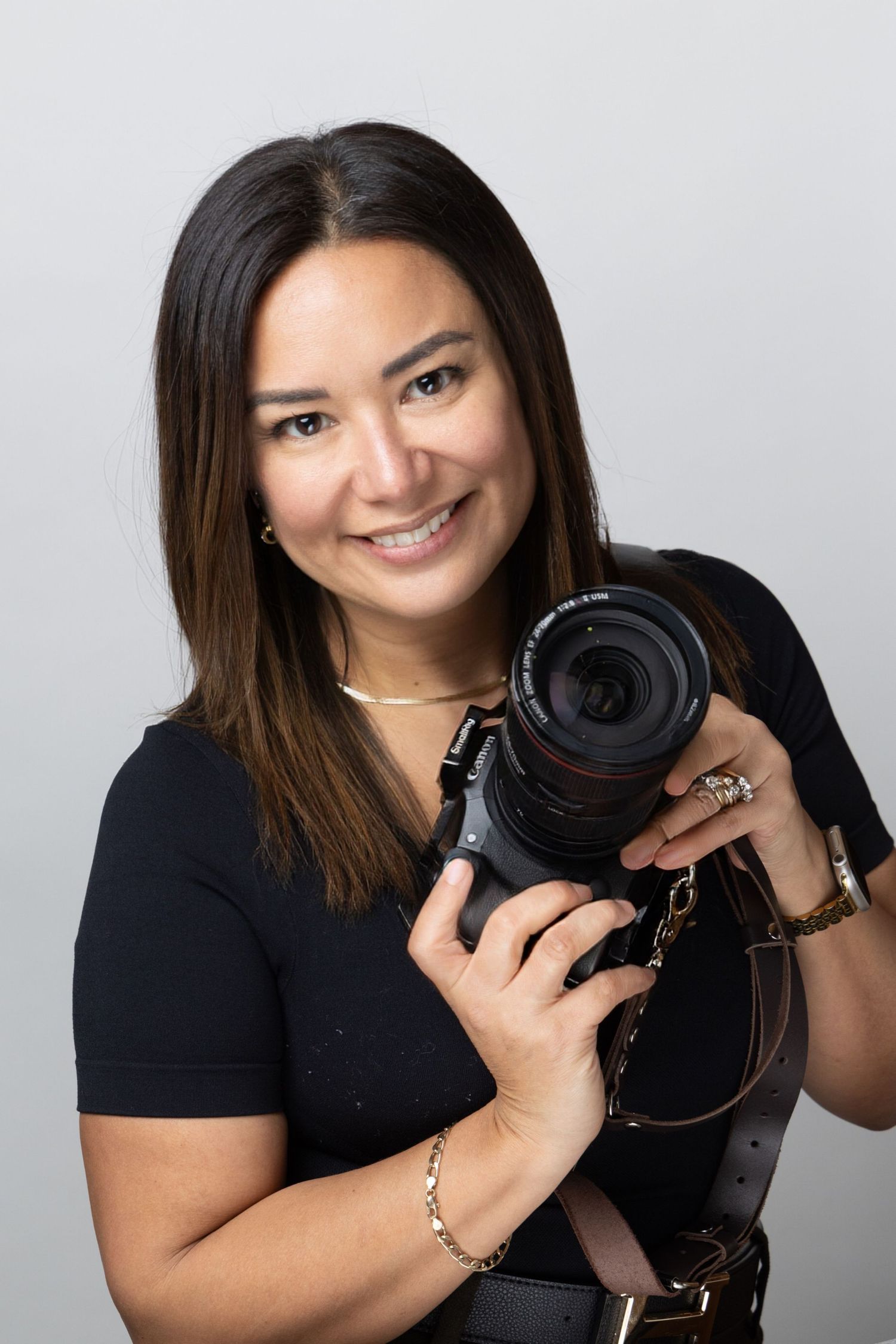 Professional photographer in black shirt poses with camera equipment against light gray studio background.