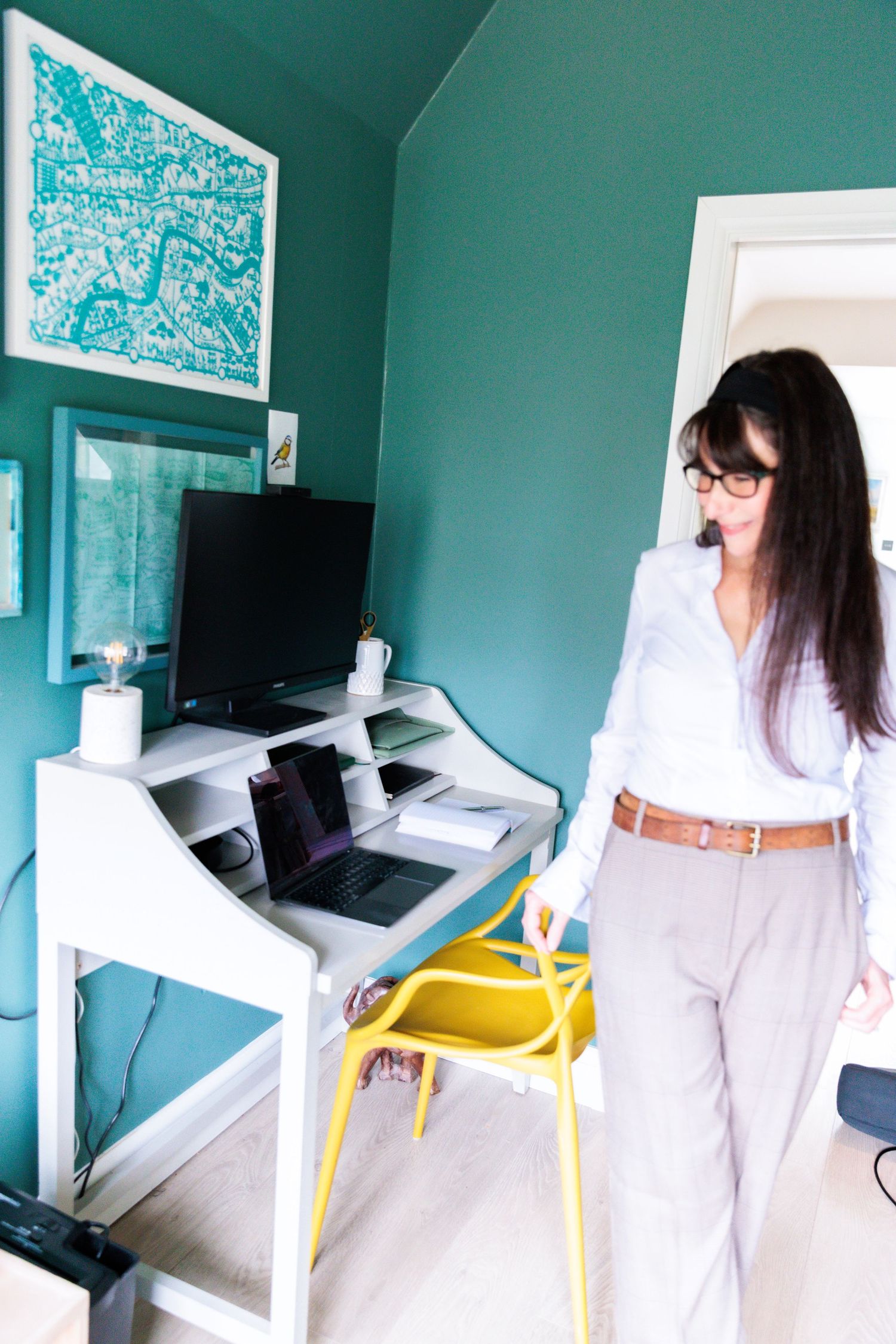 A home office setup with teal walls featuring a white desk, yellow chair, and framed artwork above the workspace.