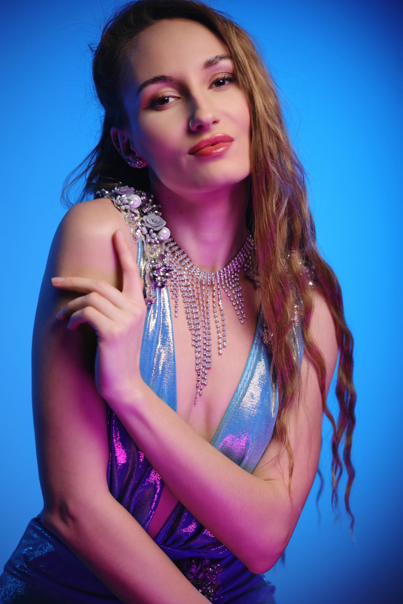 Woman in holographic outfit with crystal necklace posing against blue background under pink and purple studio lighting.