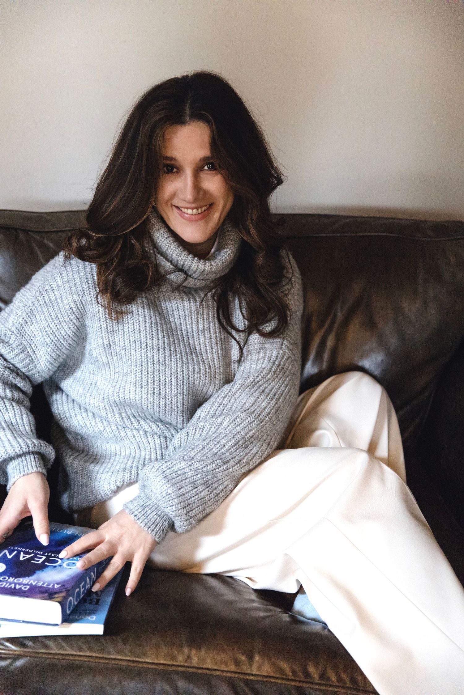 Person in gray turtleneck sweater relaxes on brown leather couch while reading a book.