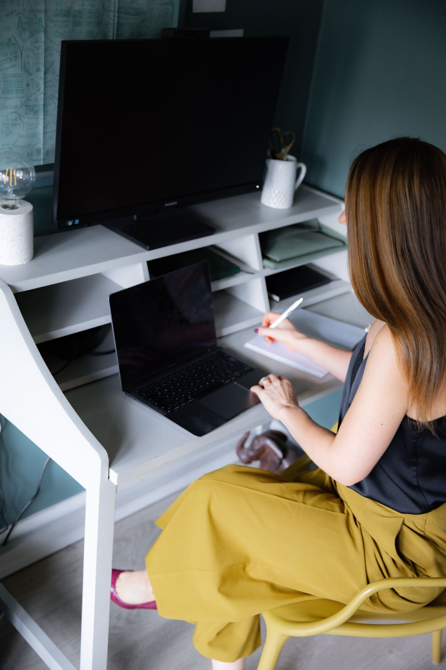 Person wearing yellow pants and black top works at white desk with laptop and monitor setup in home office space.