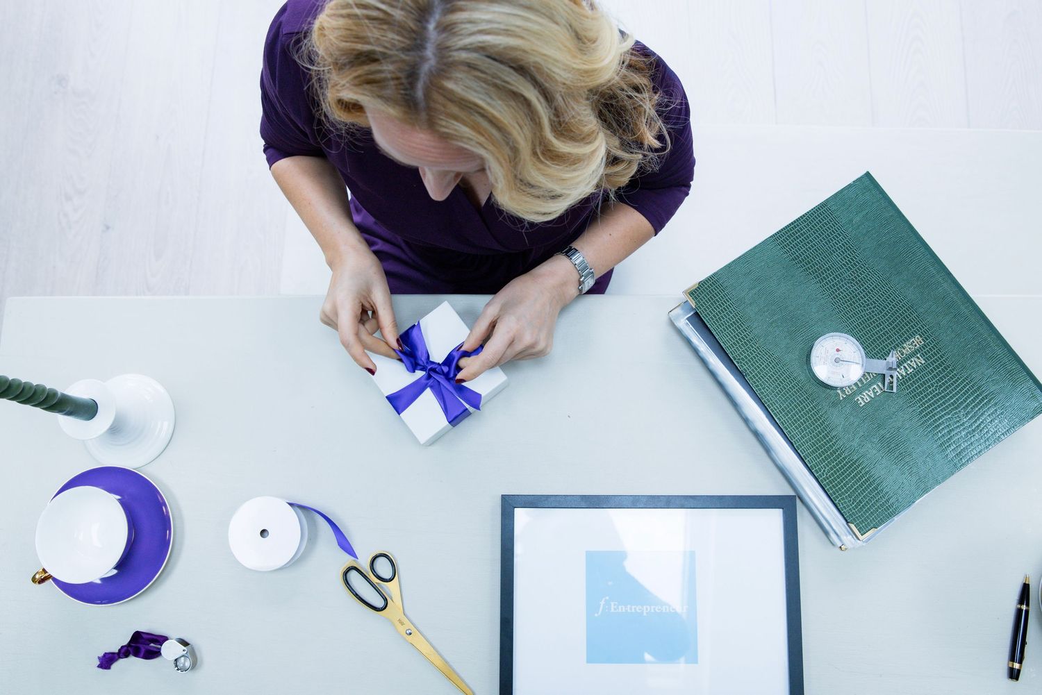 Overhead view of someone wrapping a blue and white gift with craft supplies and papers on a white surface.