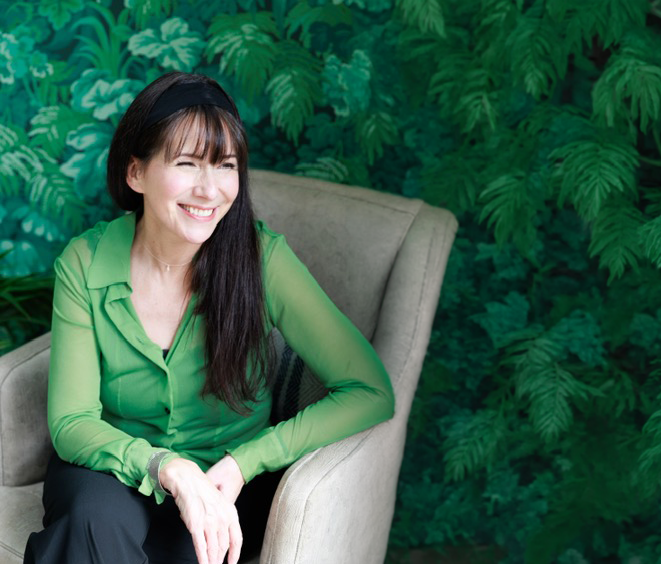 Woman in bright green button-up shirt sits in gray chair against lush green foliage background.