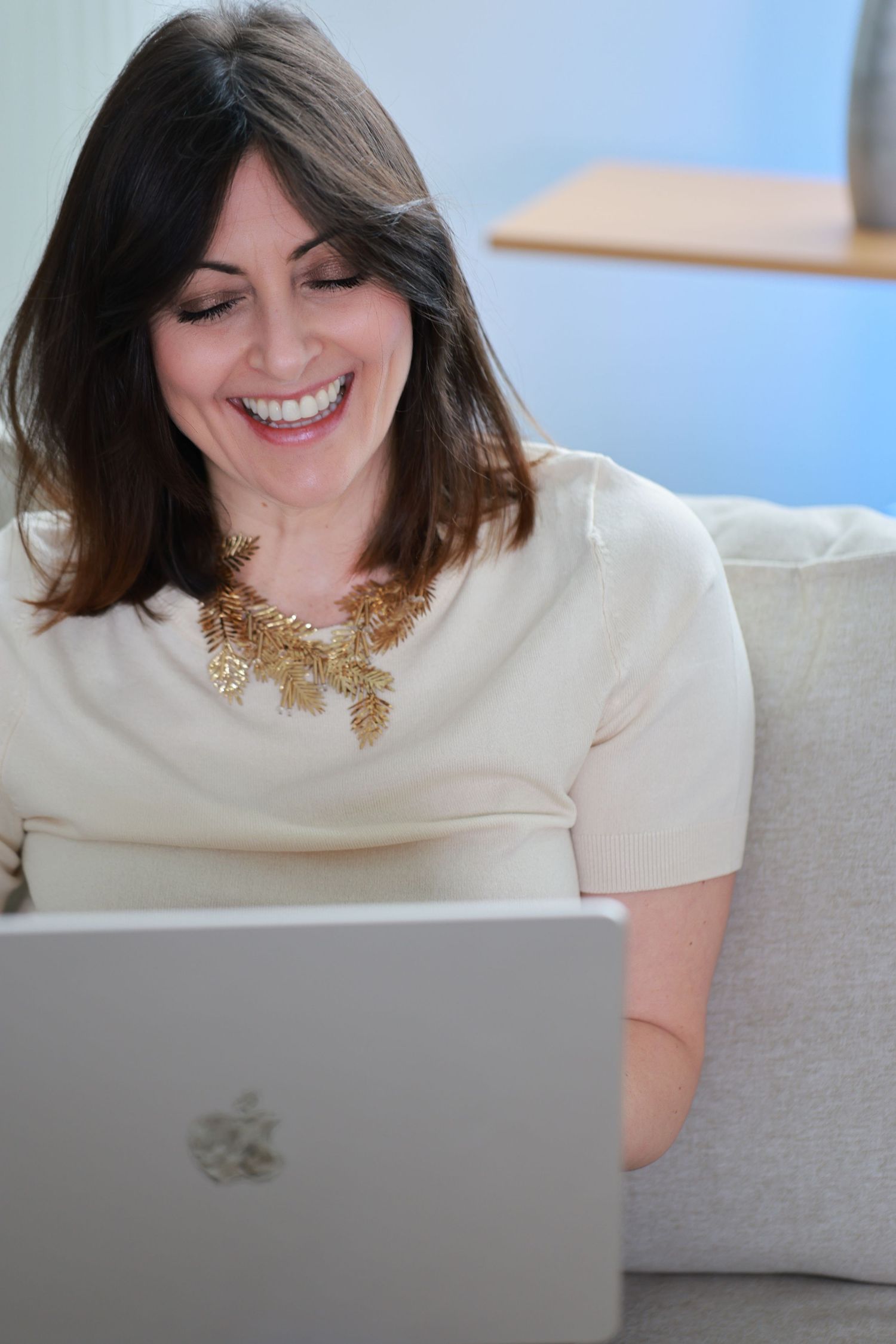 Person wearing a white shirt and gold necklace smiles while looking down at a laptop on a gray couch.
