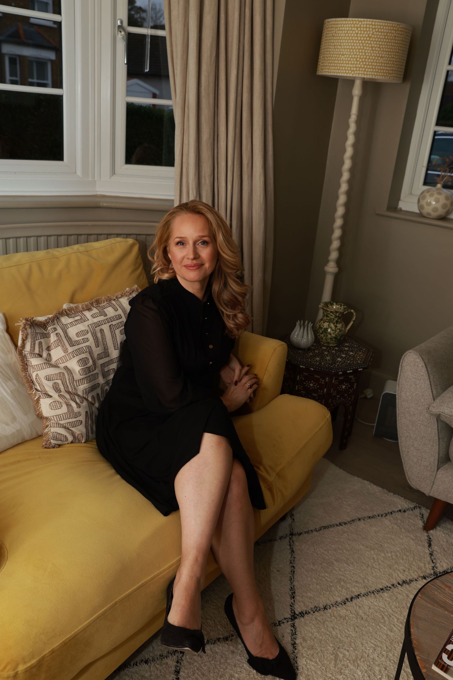 A person in a black dress sits casually on a yellow couch in a cozy living room setting.