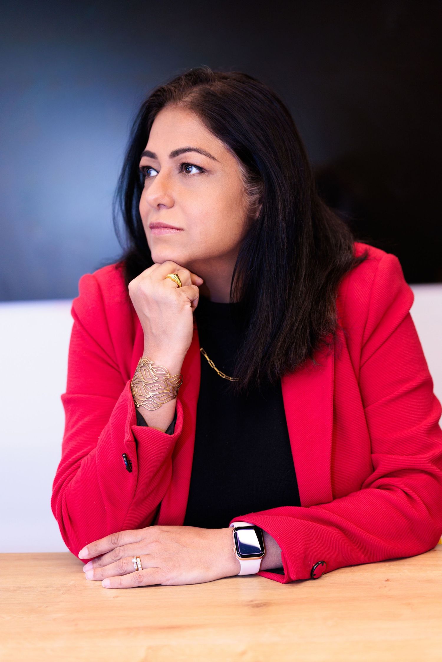 Professional woman in red blazer sitting at desk with hand on chin, wearing gold jewelry and smartwatch.