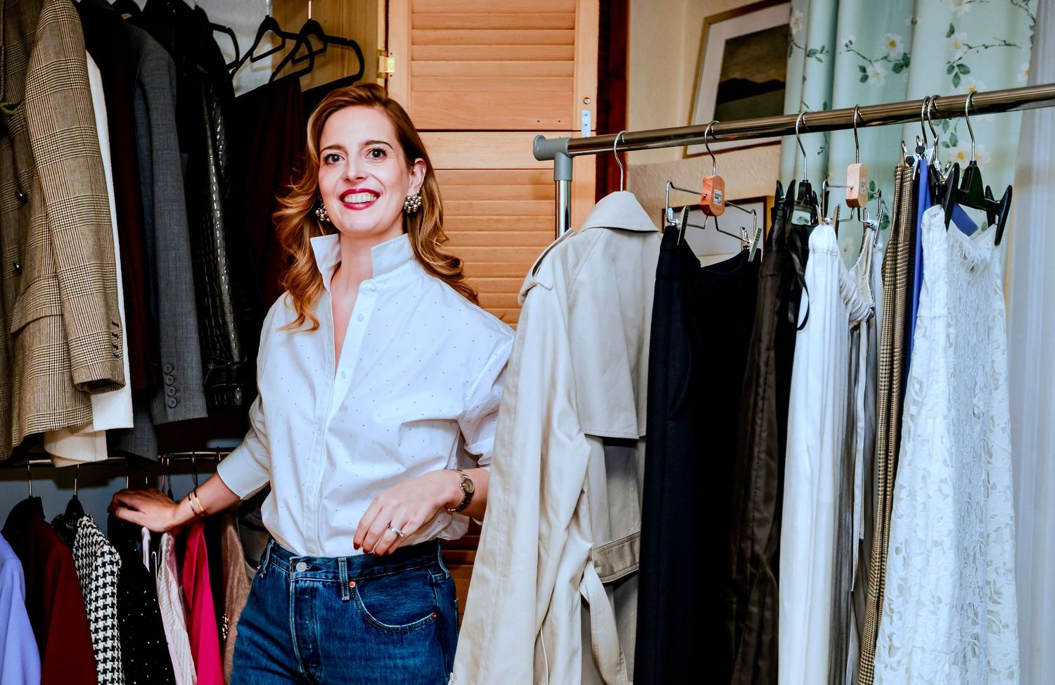 Woman smiling in a walk-in closet surrounded by hanging clothes on a rack, wearing a white shirt and jeans.