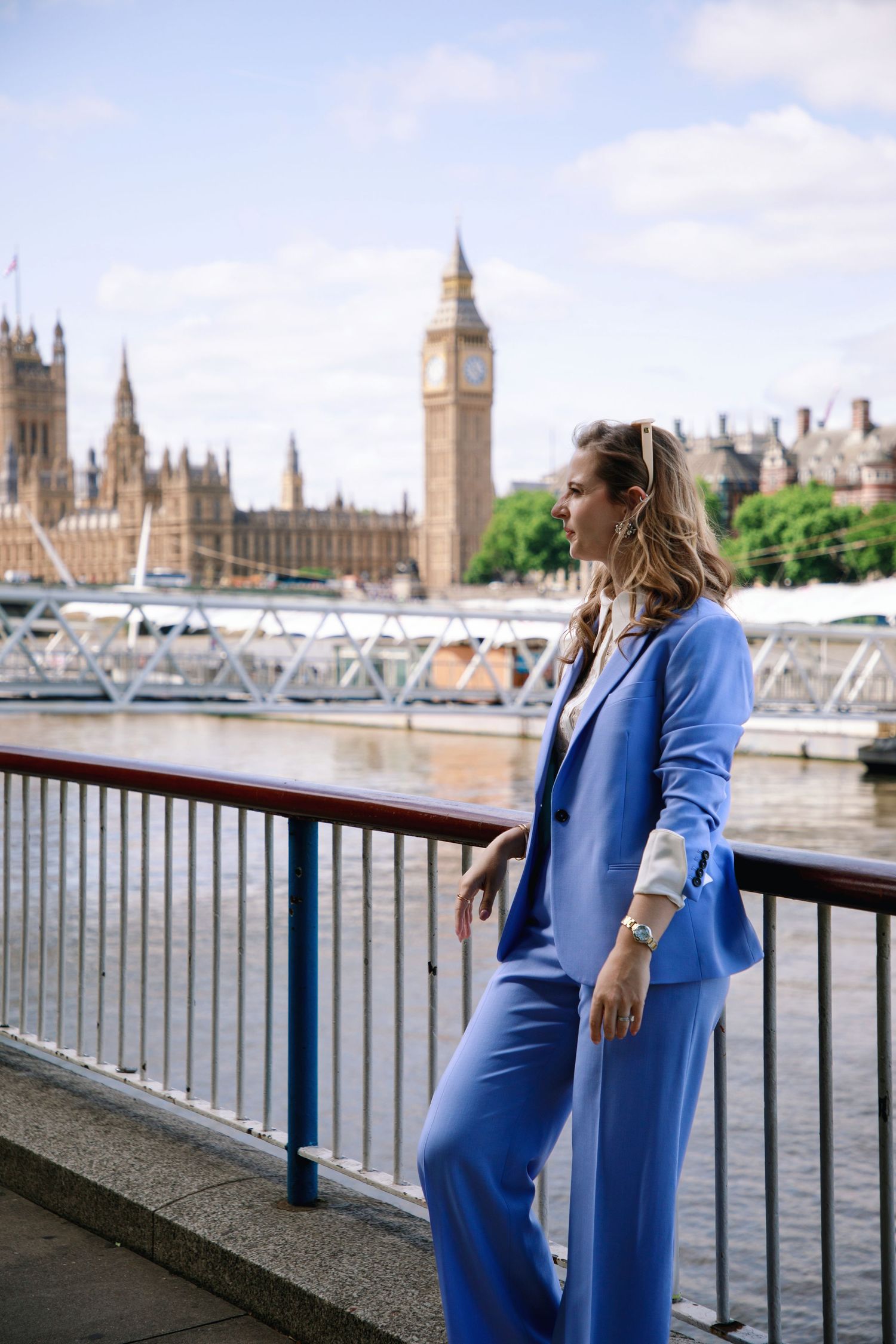 A person in a blue suit poses along the Thames River with the Houses of Parliament visible in the distance.