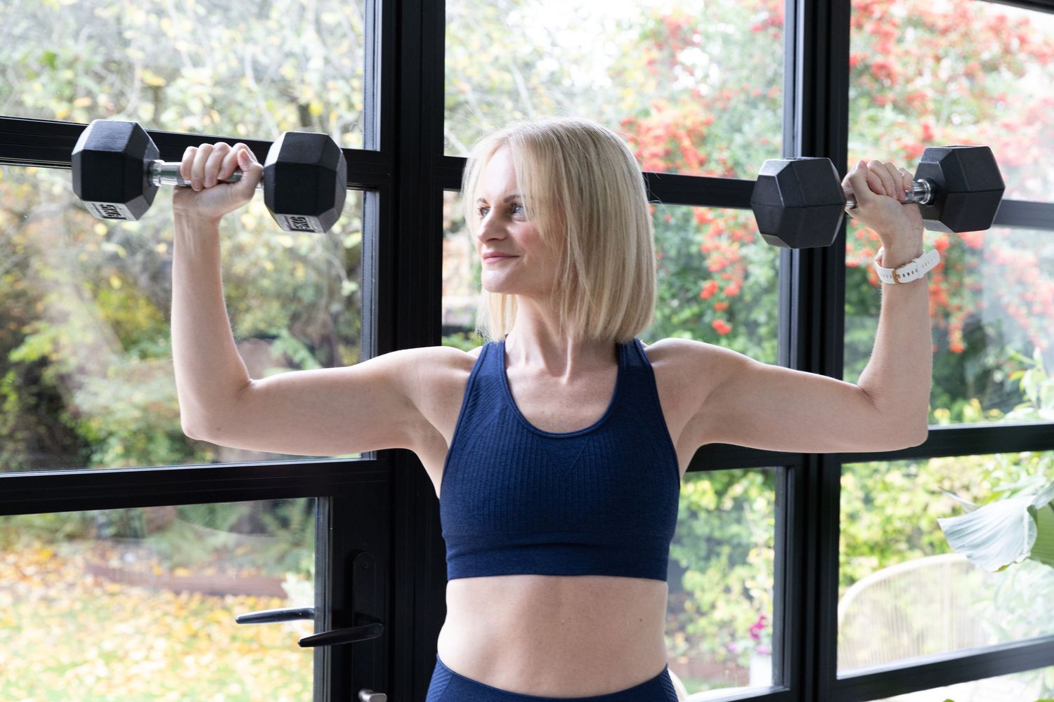 A person wearing a navy sports bra performs a cable workout routine at a fitness center with autumn foliage visible outside.
