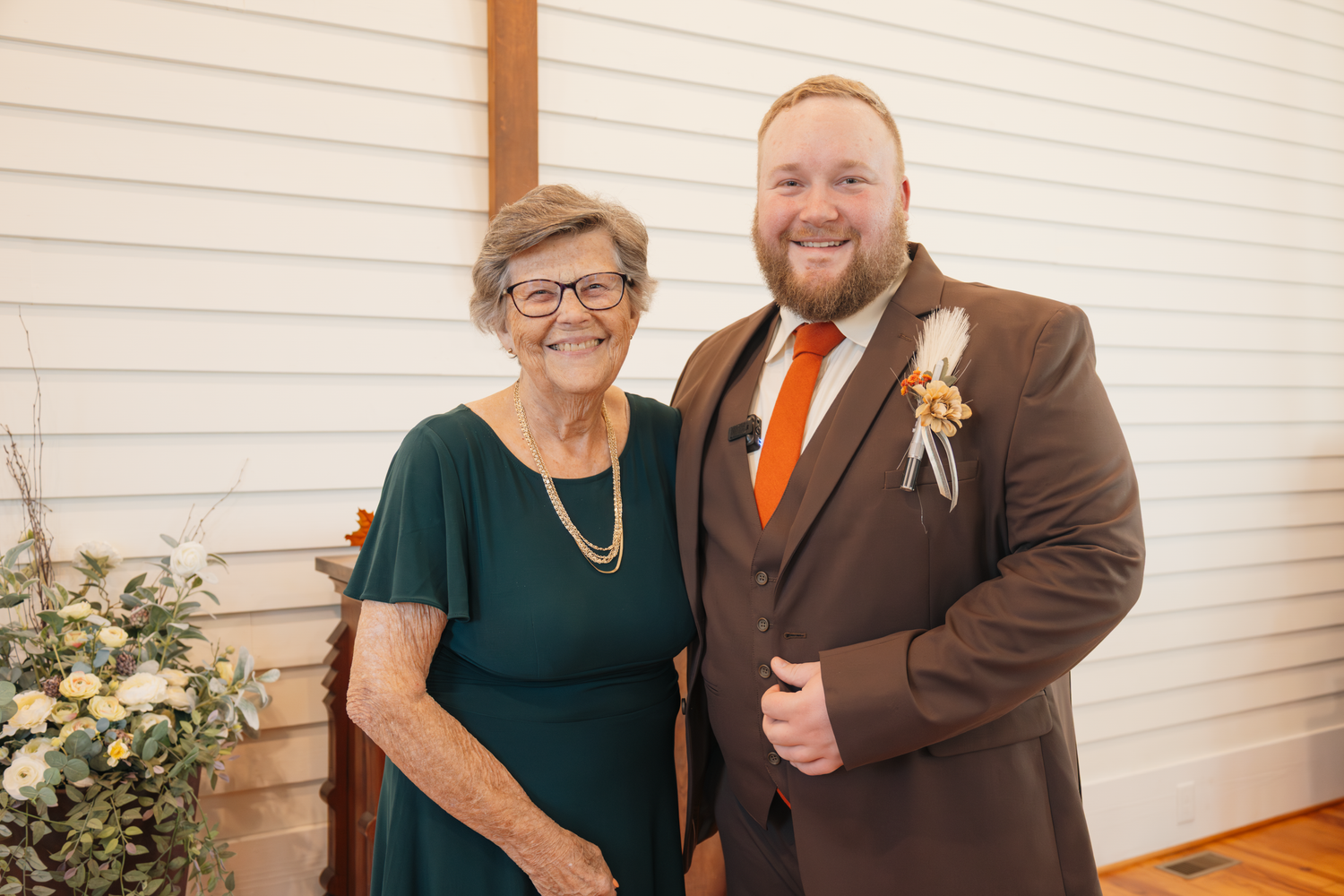 Elderly woman in green dress and young groom in brown suit with orange tie pose together at a wedding ceremony.