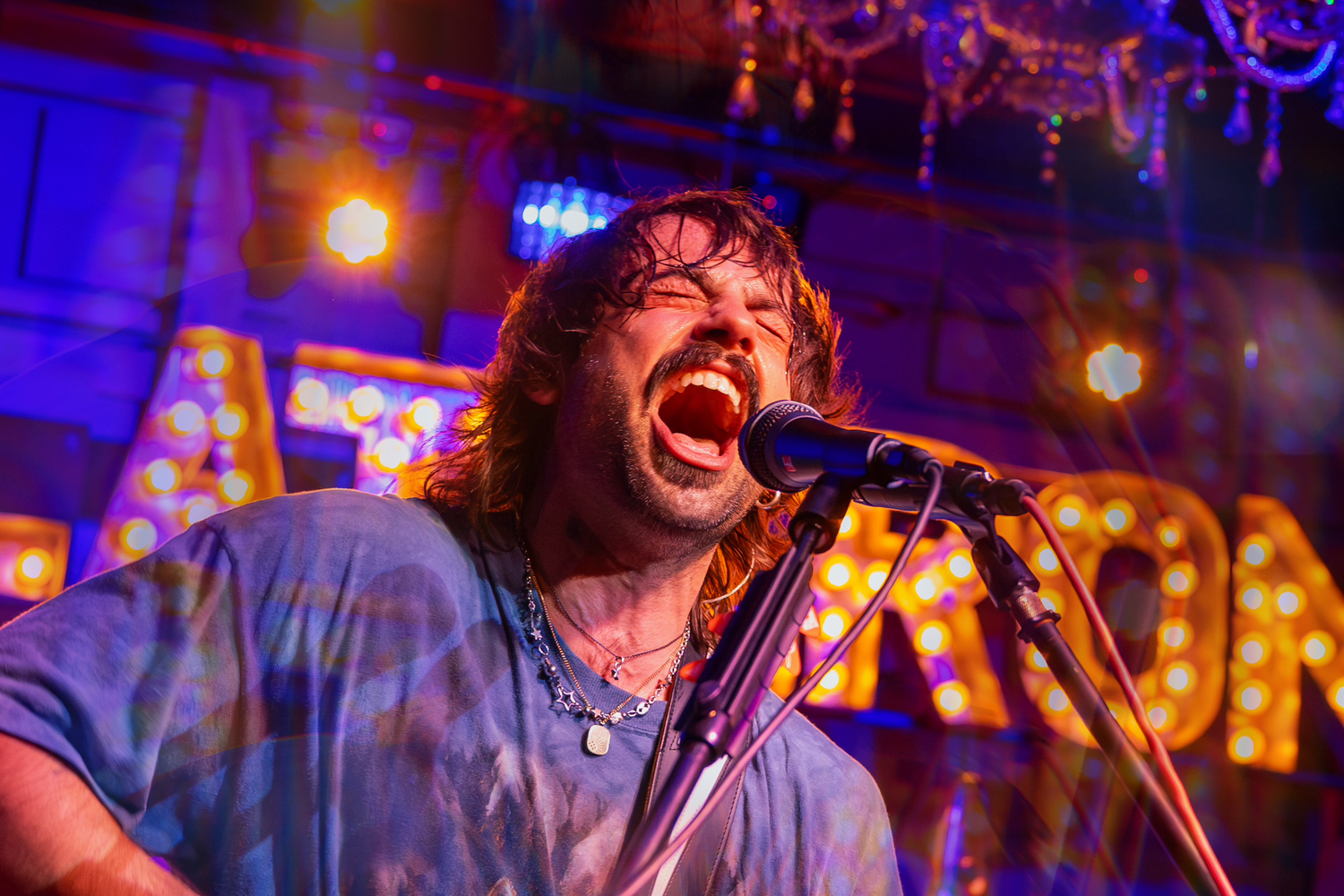 Male rock singer performing passionately on stage with colorful marquee lights in background.