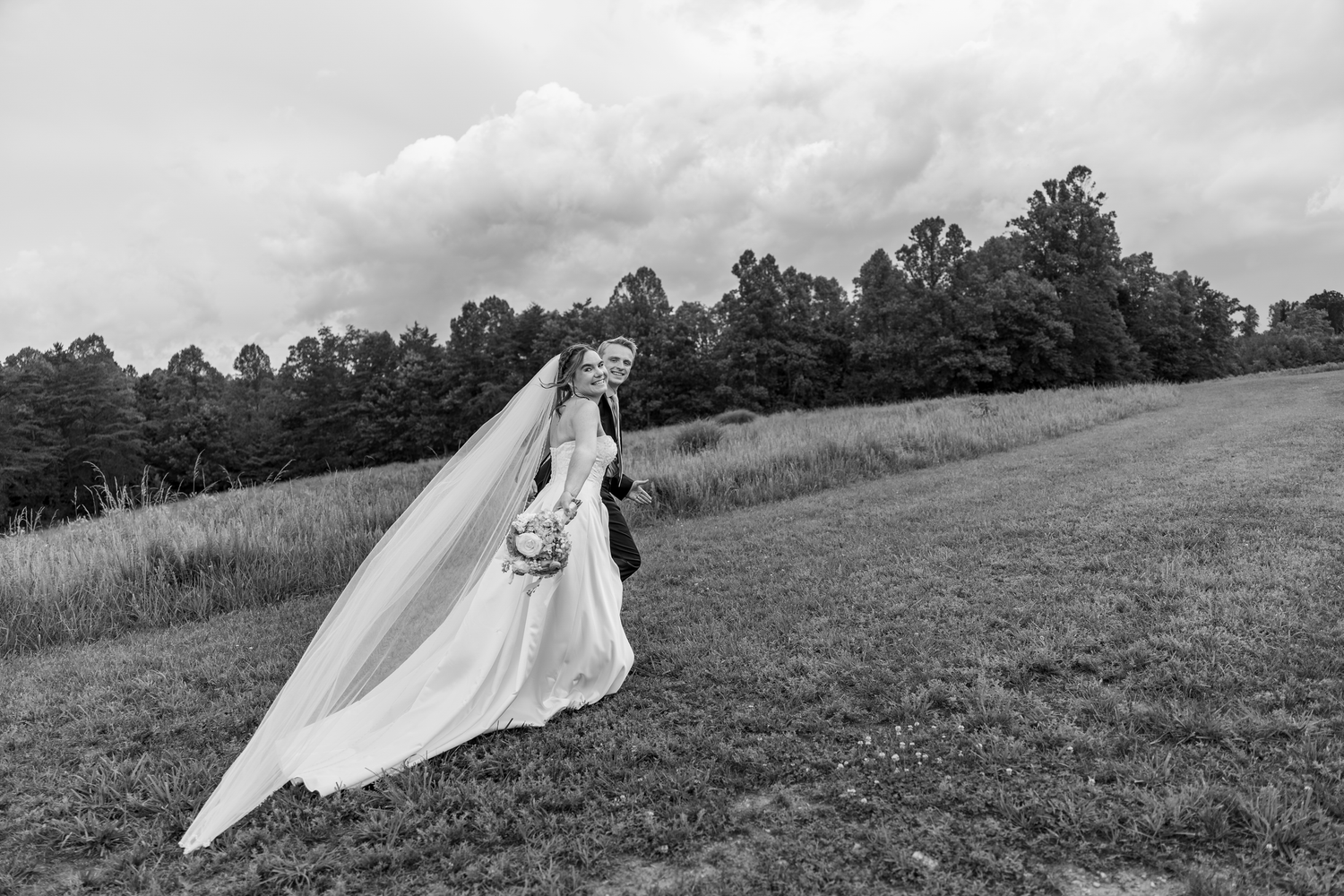 Black and white photo of a bride in a flowing gown and veil on a grassy hill with trees and cloudy sky.