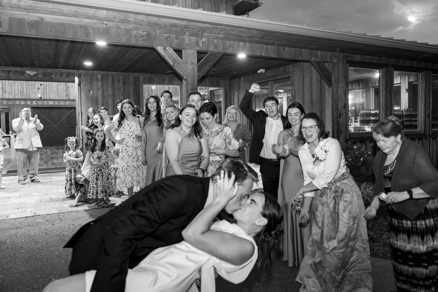 Black and white wedding reception photo with guests cheering as groom dips bride on dance floor.