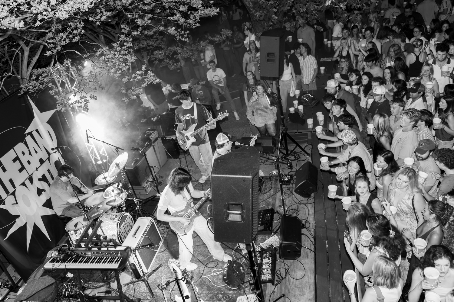 Black and white aerial view of a live band performing on stage at an outdoor concert with a large crowd.
