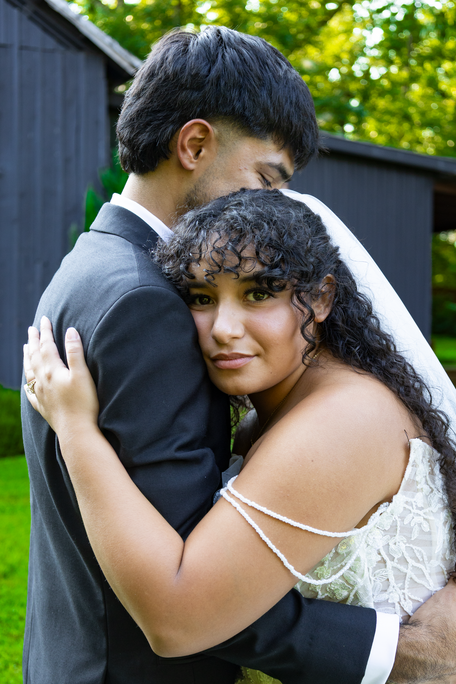 A bride in a lace gown and veil embraces her groom in a suit outdoors, sharing a tender moment on their wedding day.