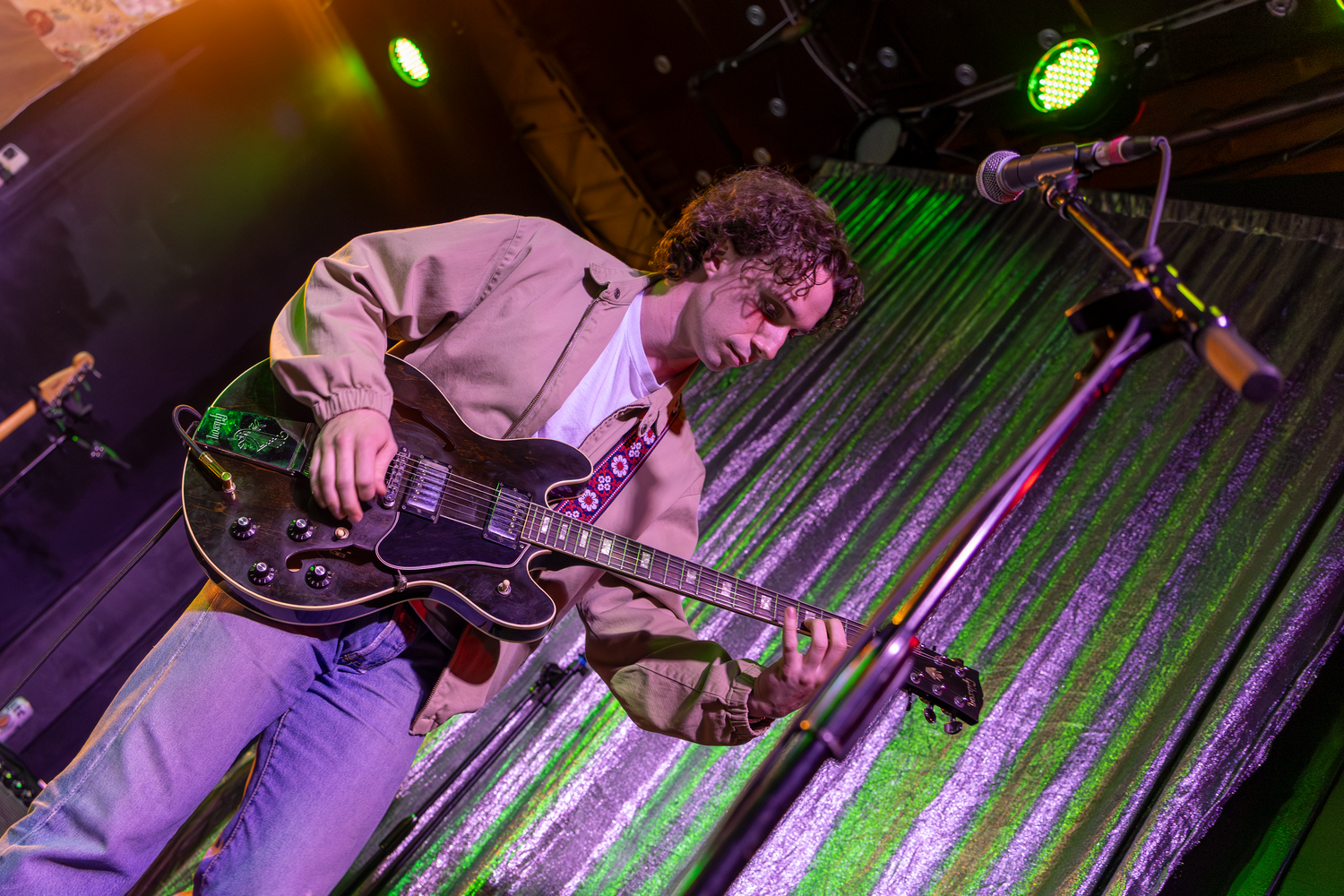 Musician performs energetically on stage with an electric guitar under colorful green and purple lighting.