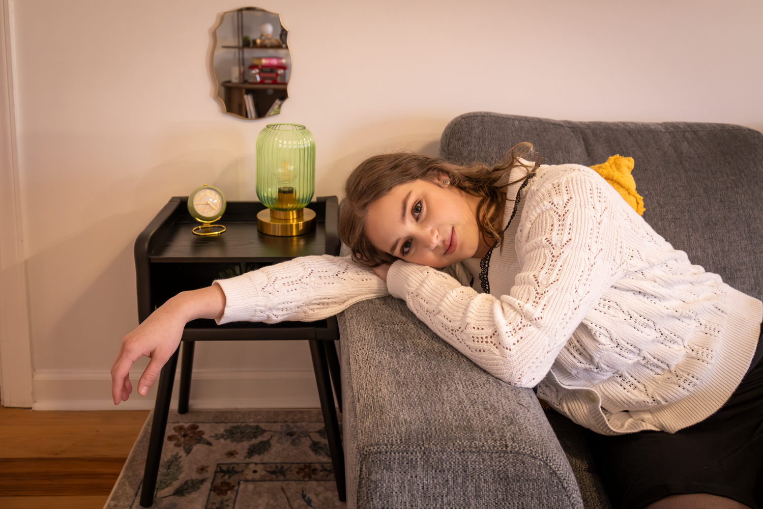 Someone rests on a gray couch next to a side table with a green lantern style lamp.