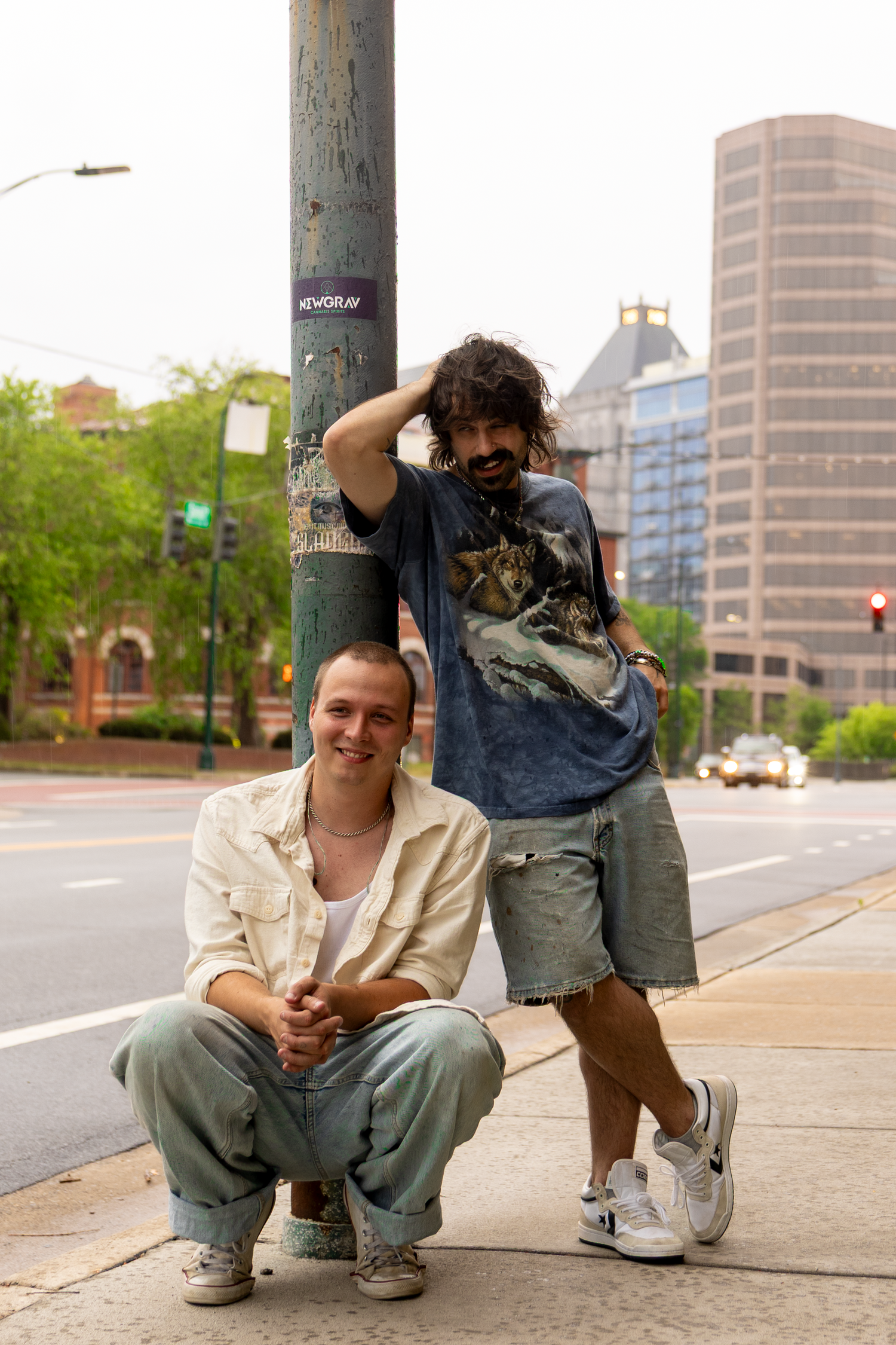 Two young men pose on a city sidewalk near a street pole, one sitting and one standing, with urban buildings in the background.
