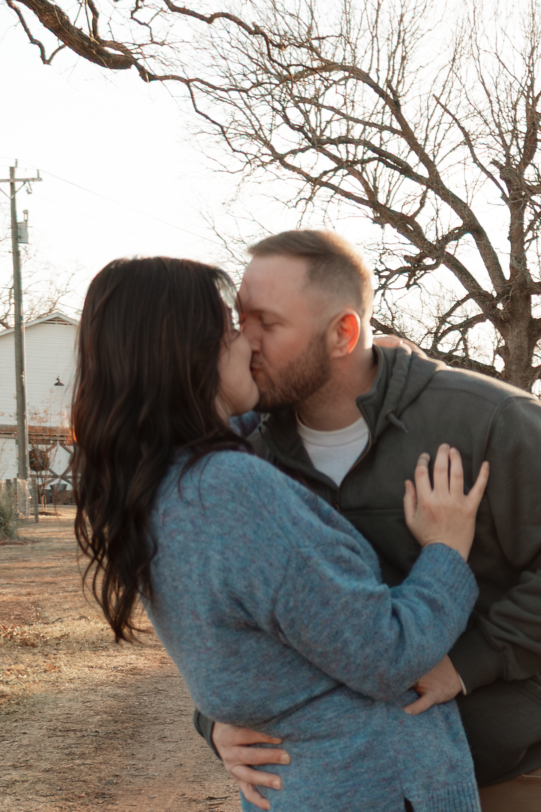 A couple shares a romantic kiss outdoors during sunset at Oak Leaf Acres Farm.