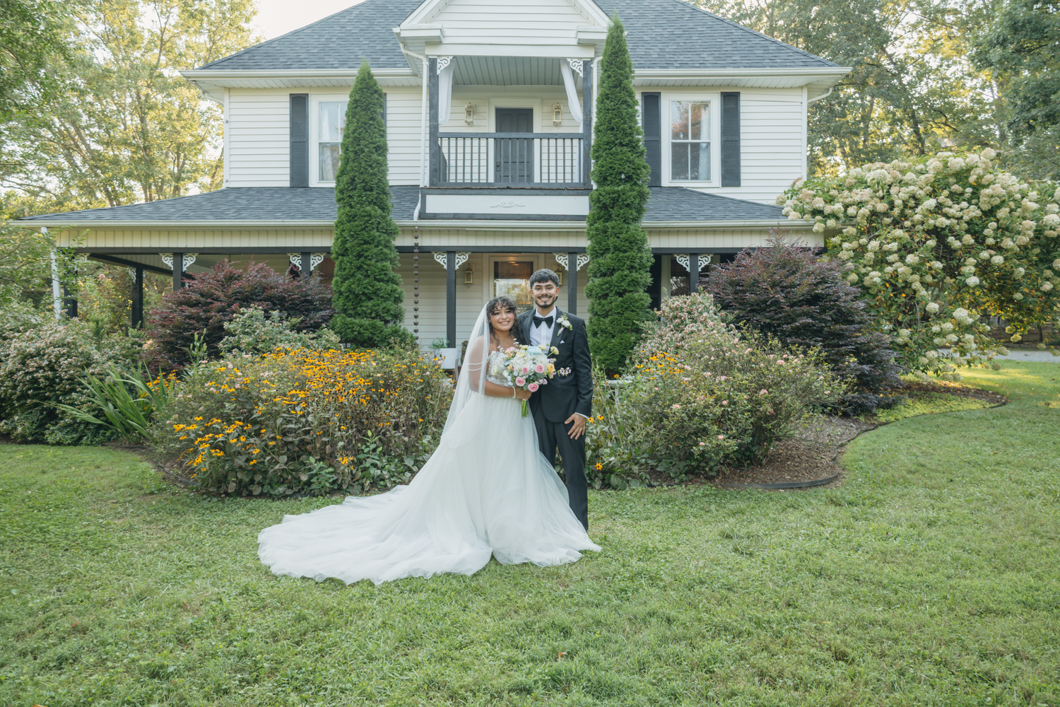 Wedding photos in front of a white house with green landscaping and cypress trees at The Farmhouse Siloam.