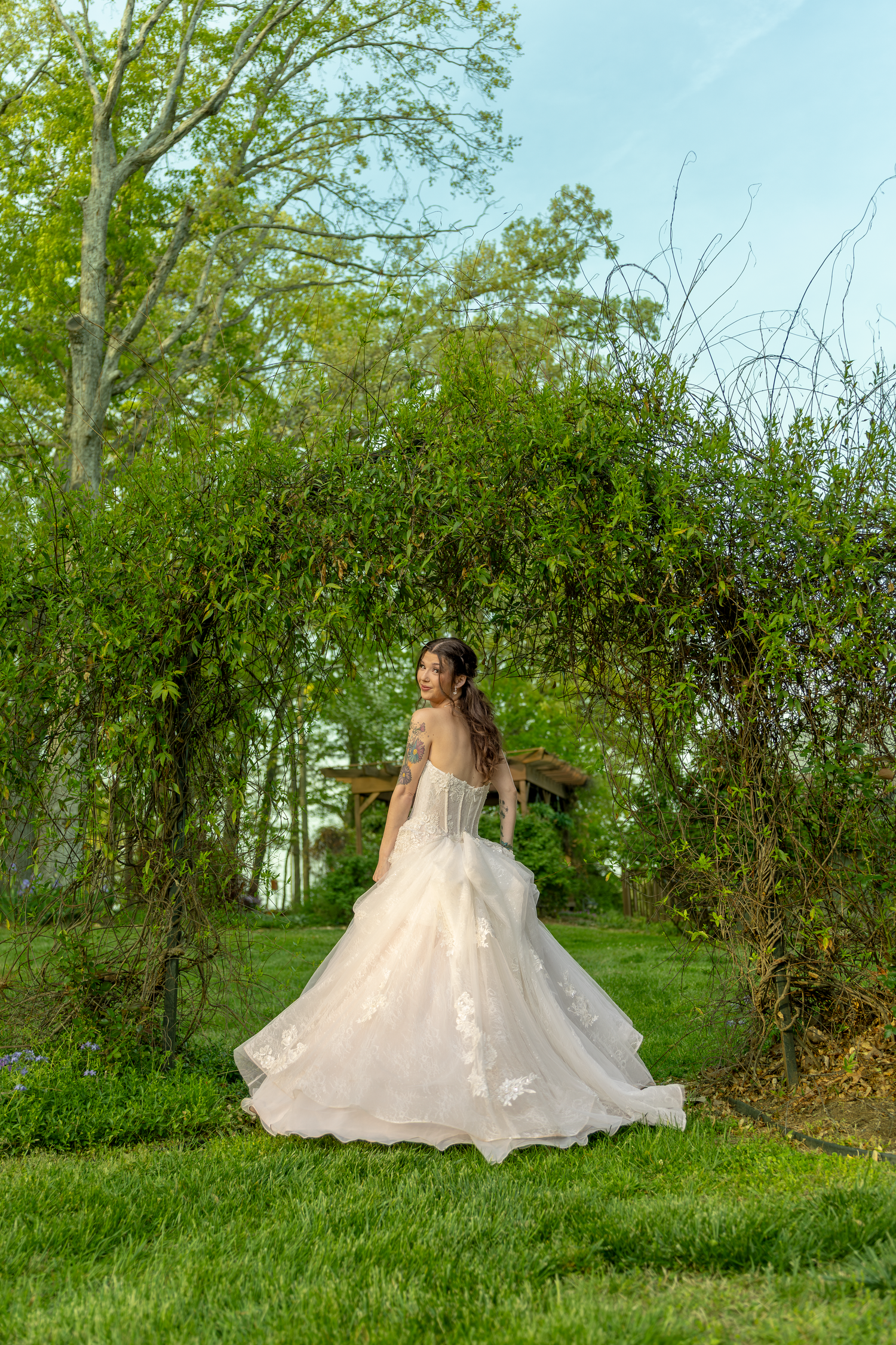 Bride in flowing white ball gown twirling under a lush green garden arch on a sunny day.