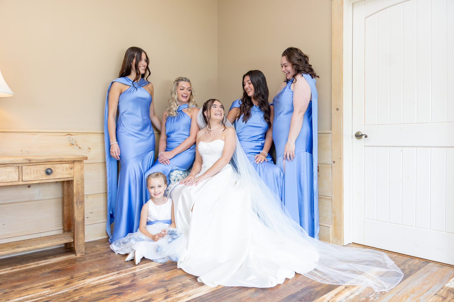 Bride in white gown sits with flower girl surrounded by bridesmaids in blue dresses in elegant room.