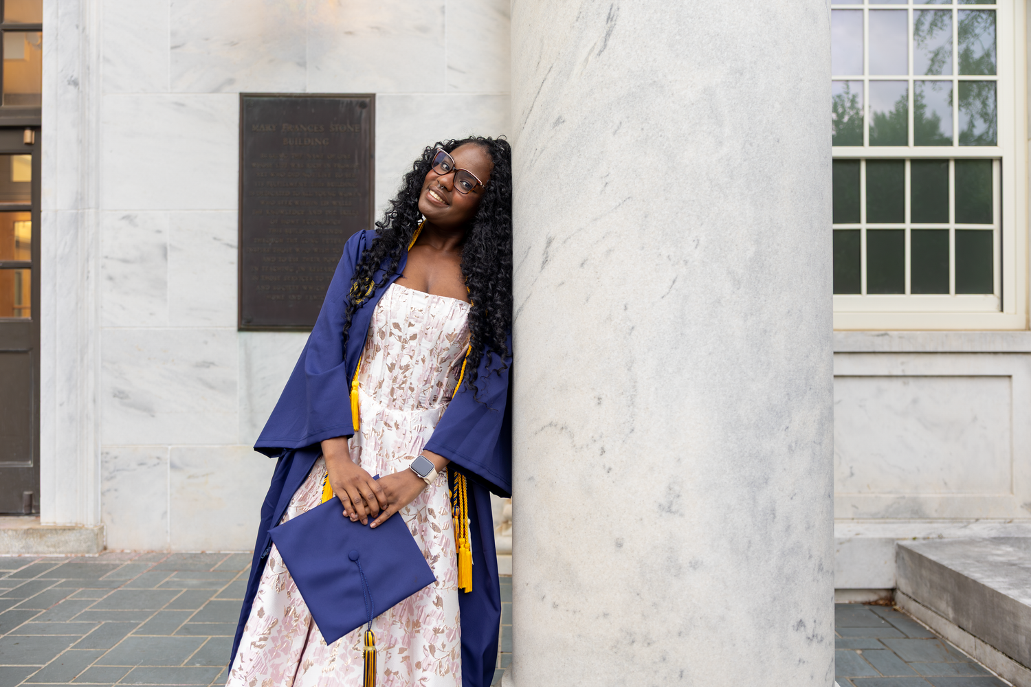 Young woman in navy graduation gown and cap leans against marble column outdoors, wearing white floral dress.