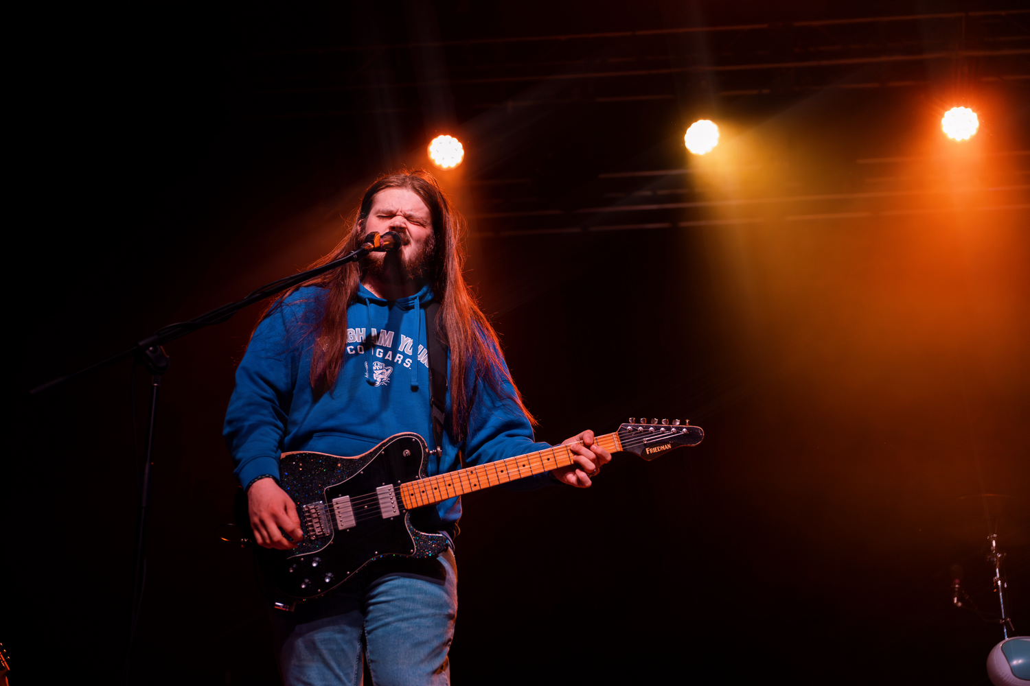 Musicians perform on stage with orange lighting and guitar during a live concert performance.