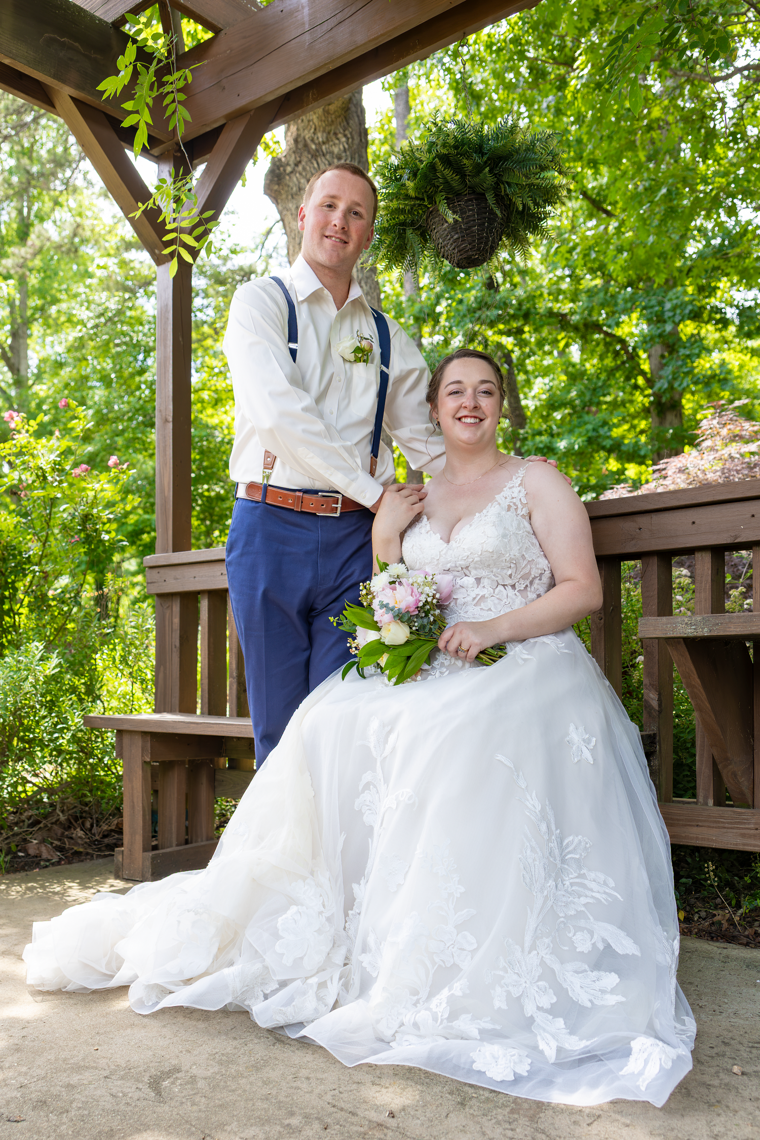 Bride in white gown holding bouquet sits beside groom in blue suit under wooden gazebo surrounded by lush greenery.