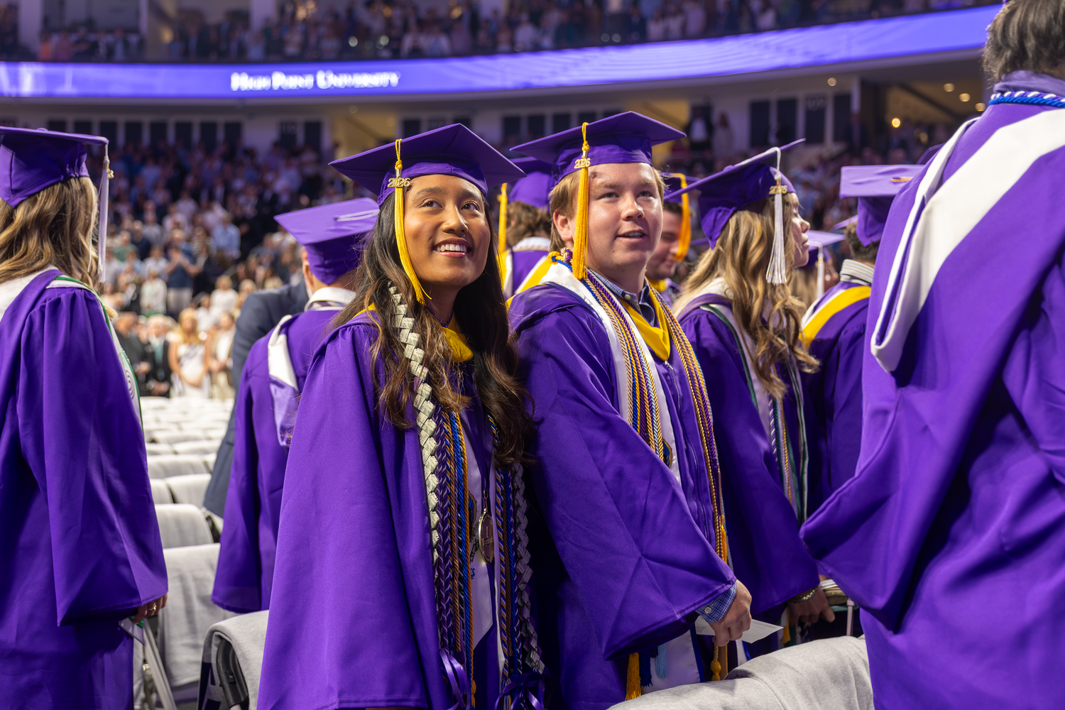 Students in purple graduation caps and gowns smile during a university commencement ceremony indoors.