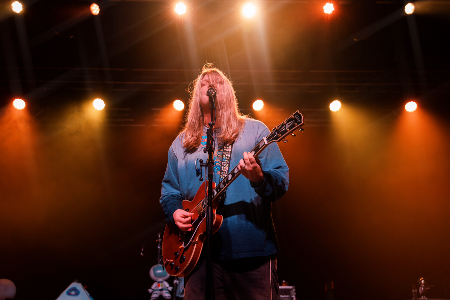Concert performers play guitars on stage under warm orange spotlights during a live music performance.