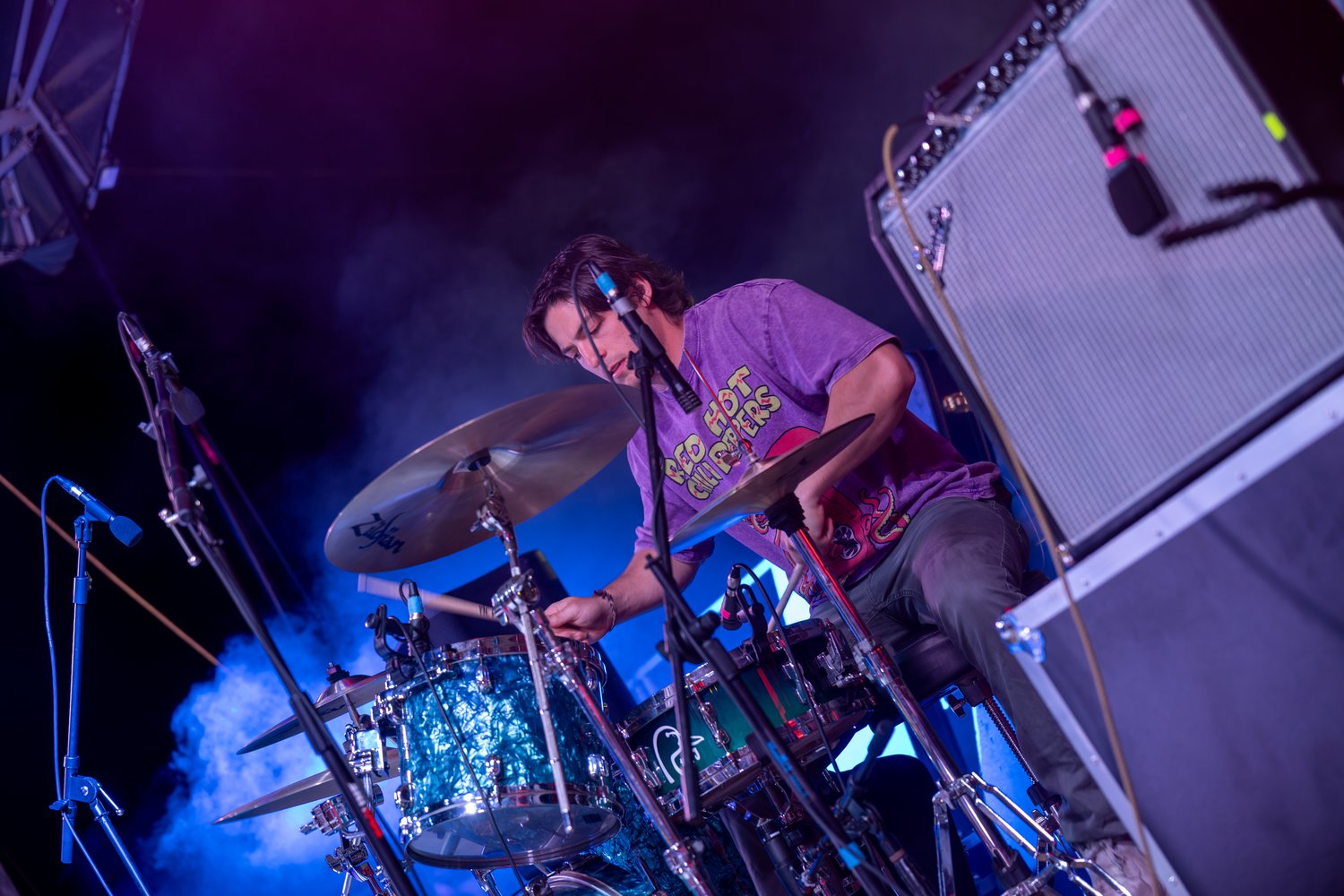 A drummer performing live on stage under purple lights, playing cymbals with energy at an outdoor music event.