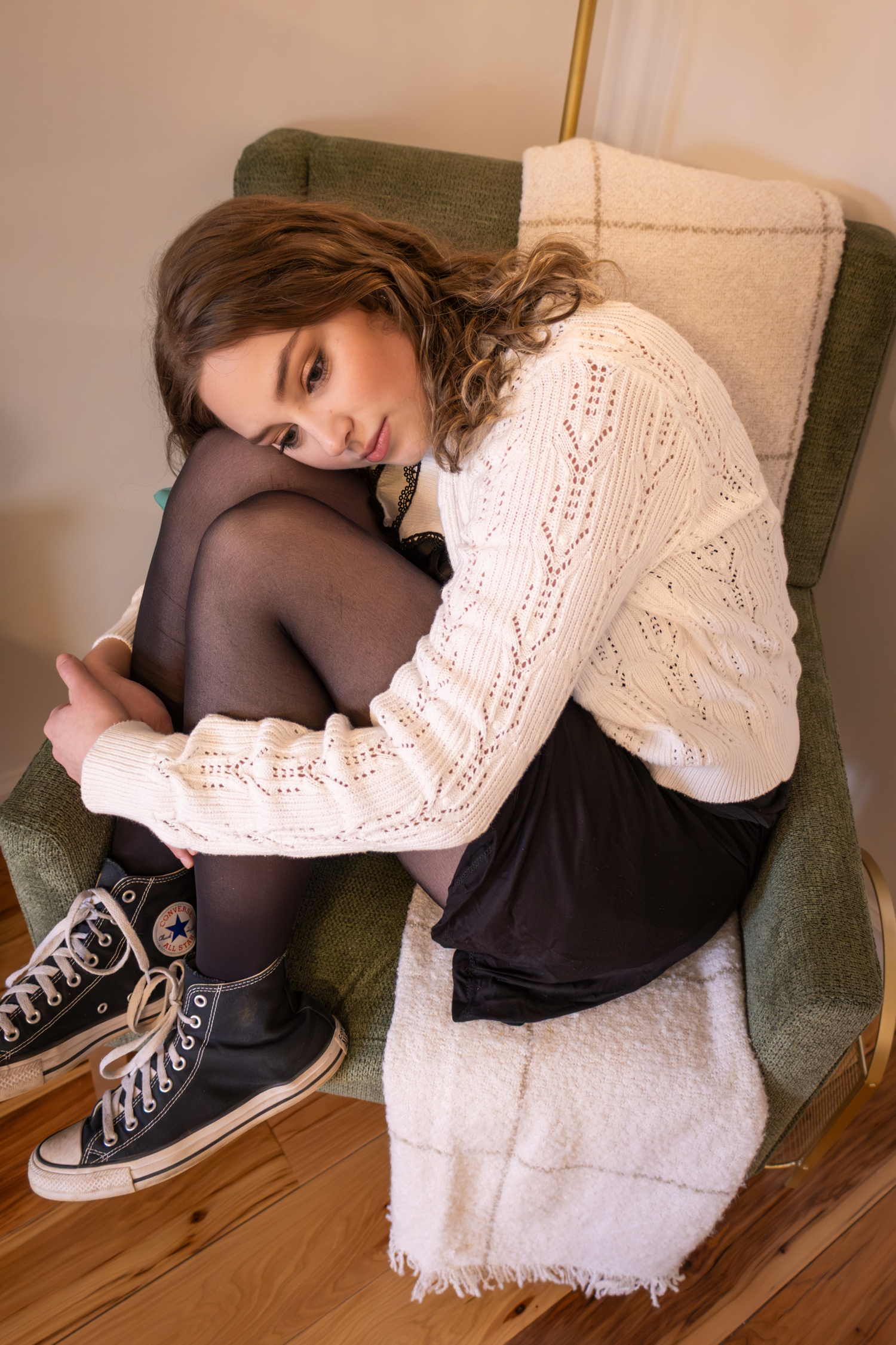 A person in a white knit sweater and black tights sits curled up in a green armchair.