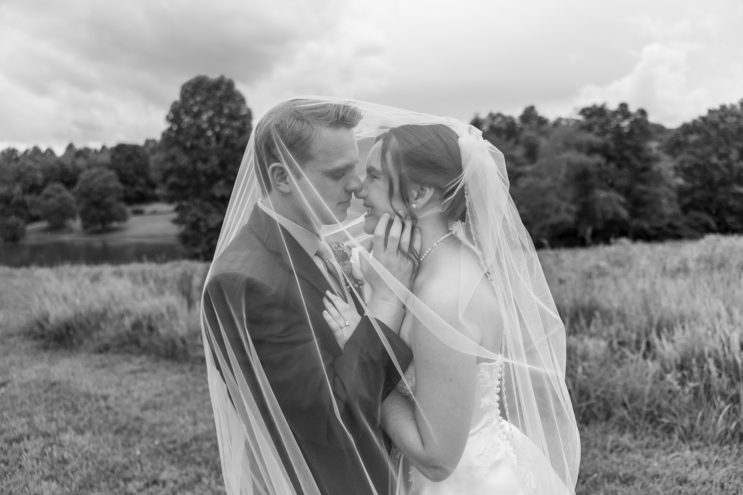 Black and white photo of bride and groom sharing intimate moment under veil in lush outdoor setting.