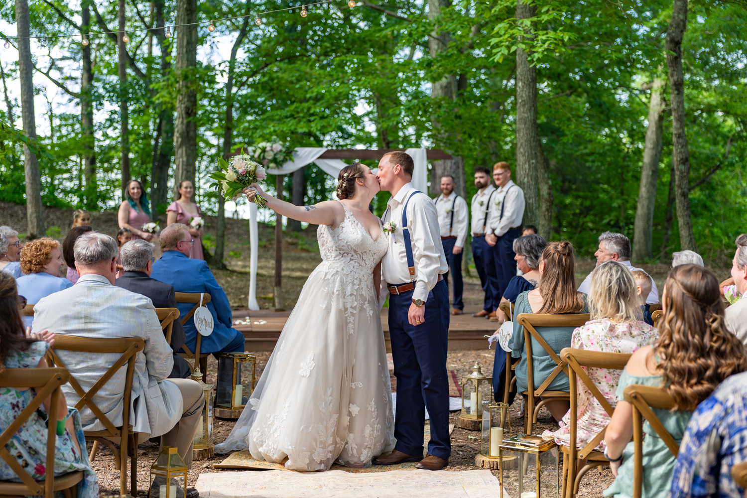 Bride and groom walk down outdoor wedding aisle surrounded by guests and tall trees in a romantic forest ceremony.