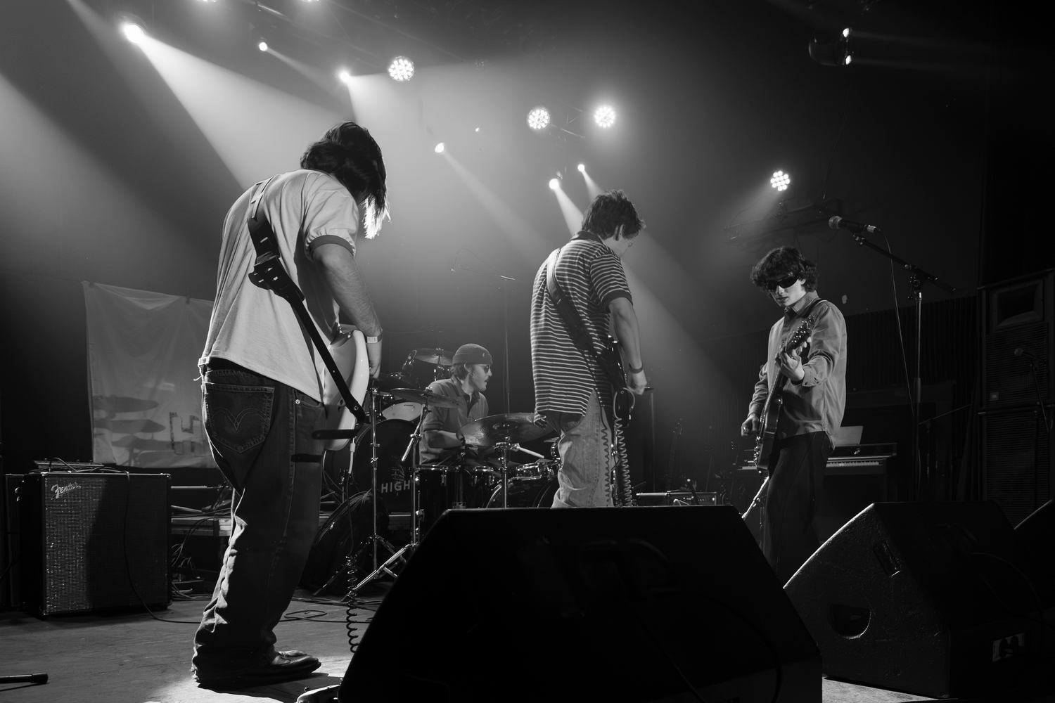 Black and white concert photography showing musicians performing on a darkened stage with spotlights.