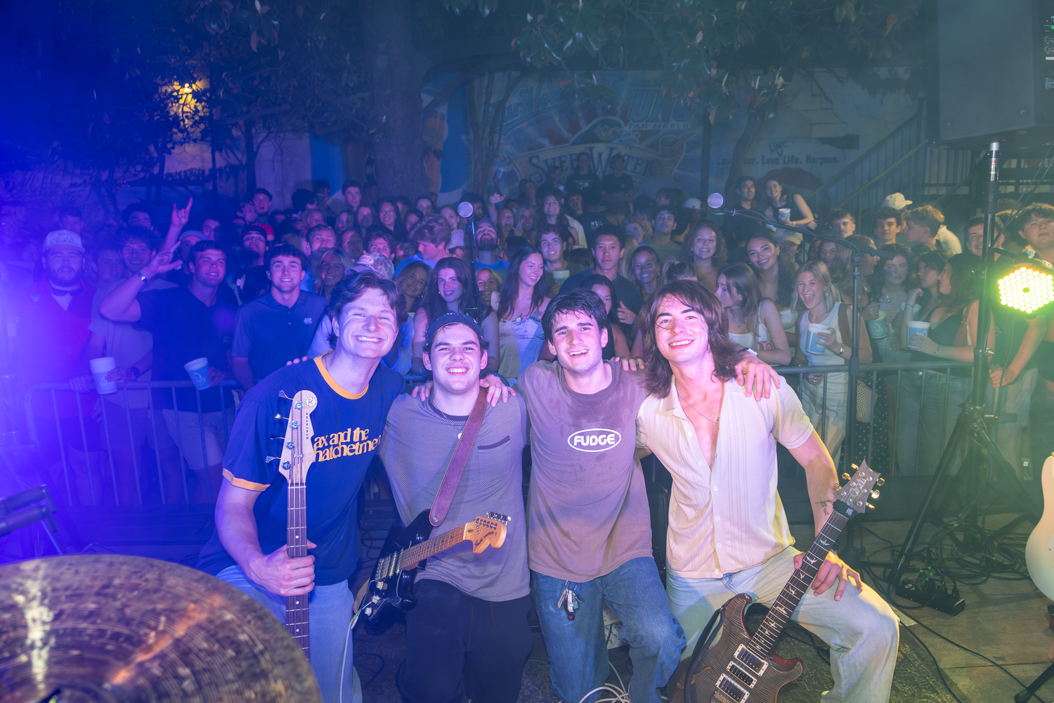 Four young band members pose together smiling in front of a large crowd at an outdoor concert at night.