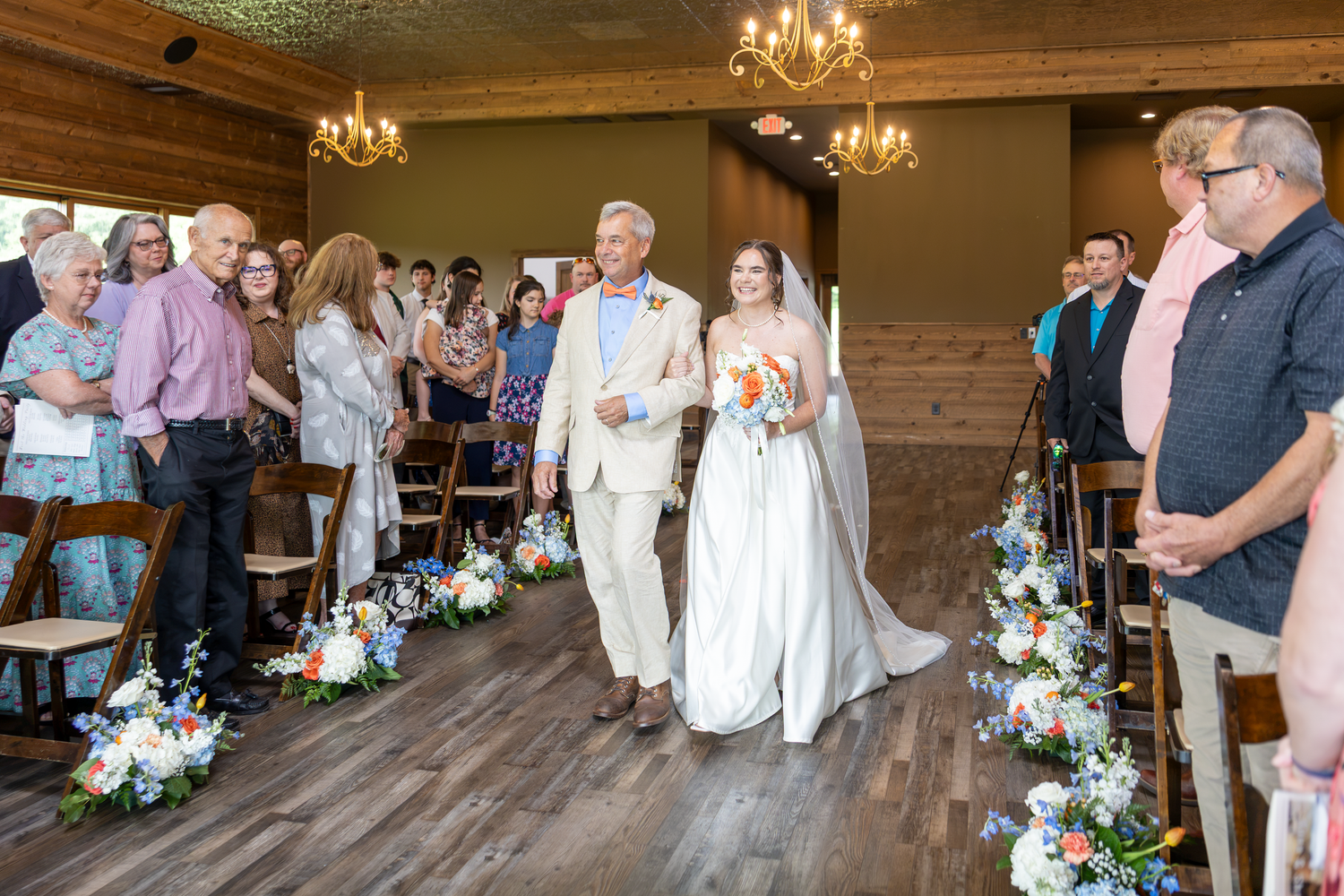 Bride walks down the aisle with her father at an elegant indoor wedding ceremony with floral arrangements.
