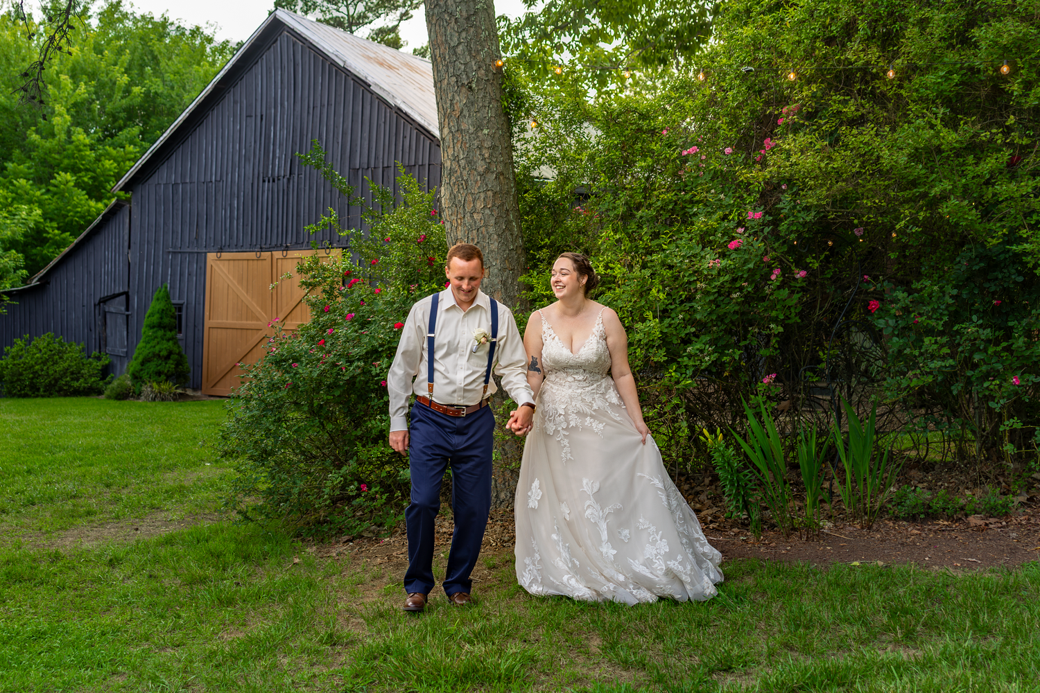 Bride and groom walk hand in hand on lush green lawn beside rustic dark barn surrounded by trees.